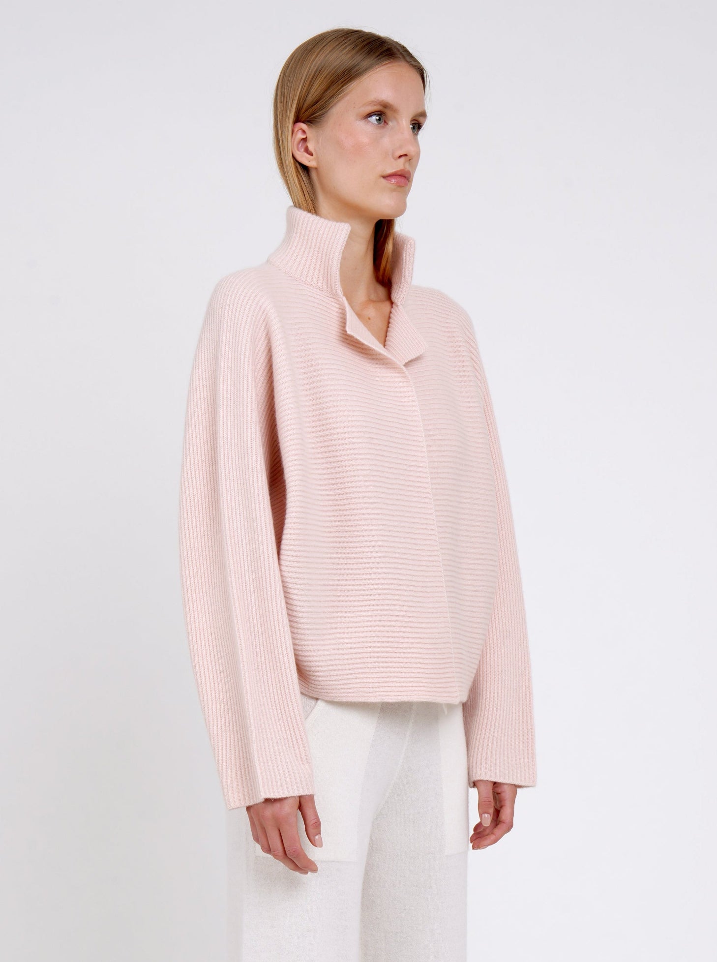 Side view close-up modal in relaxed fit ribbed knit peach cashmere jacket with high collar kimono sleeves and white wide leg knit pants.