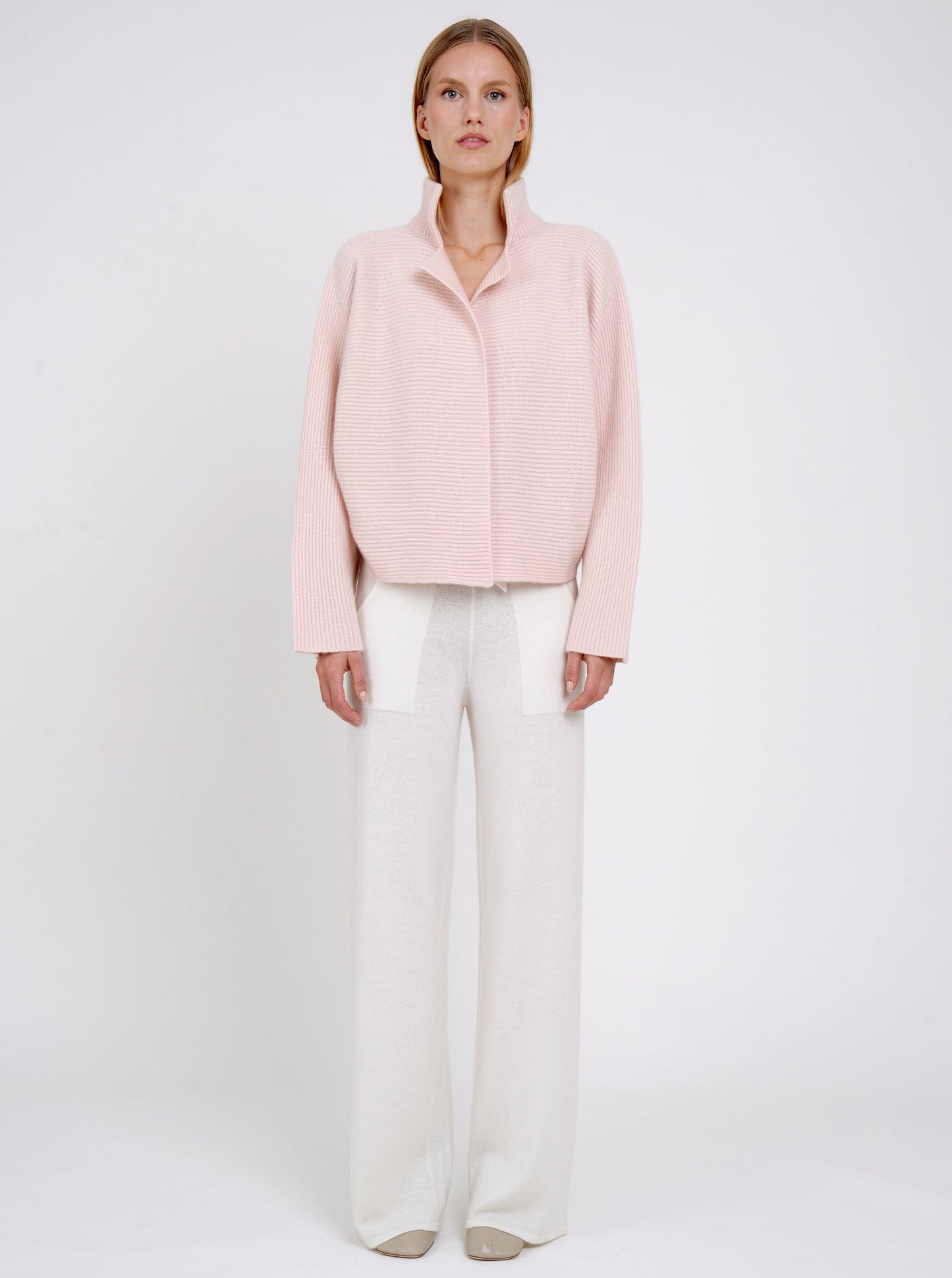 Front view modal in ribbed knit peach cashmere short jacket with high collar kimono sleeves and white wide leg knit pants.