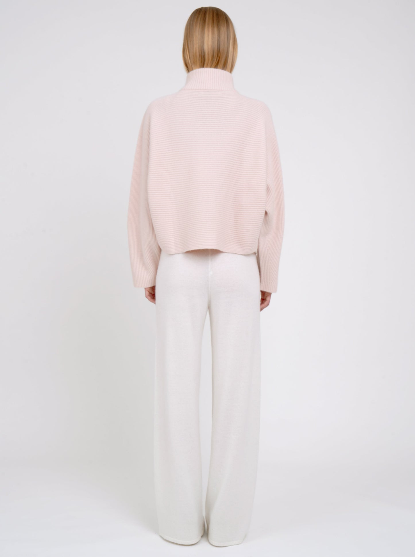 Back view modal in ribbed knit peach cashmere short jacket with high collar kimono sleeves and white wide leg knit pants.