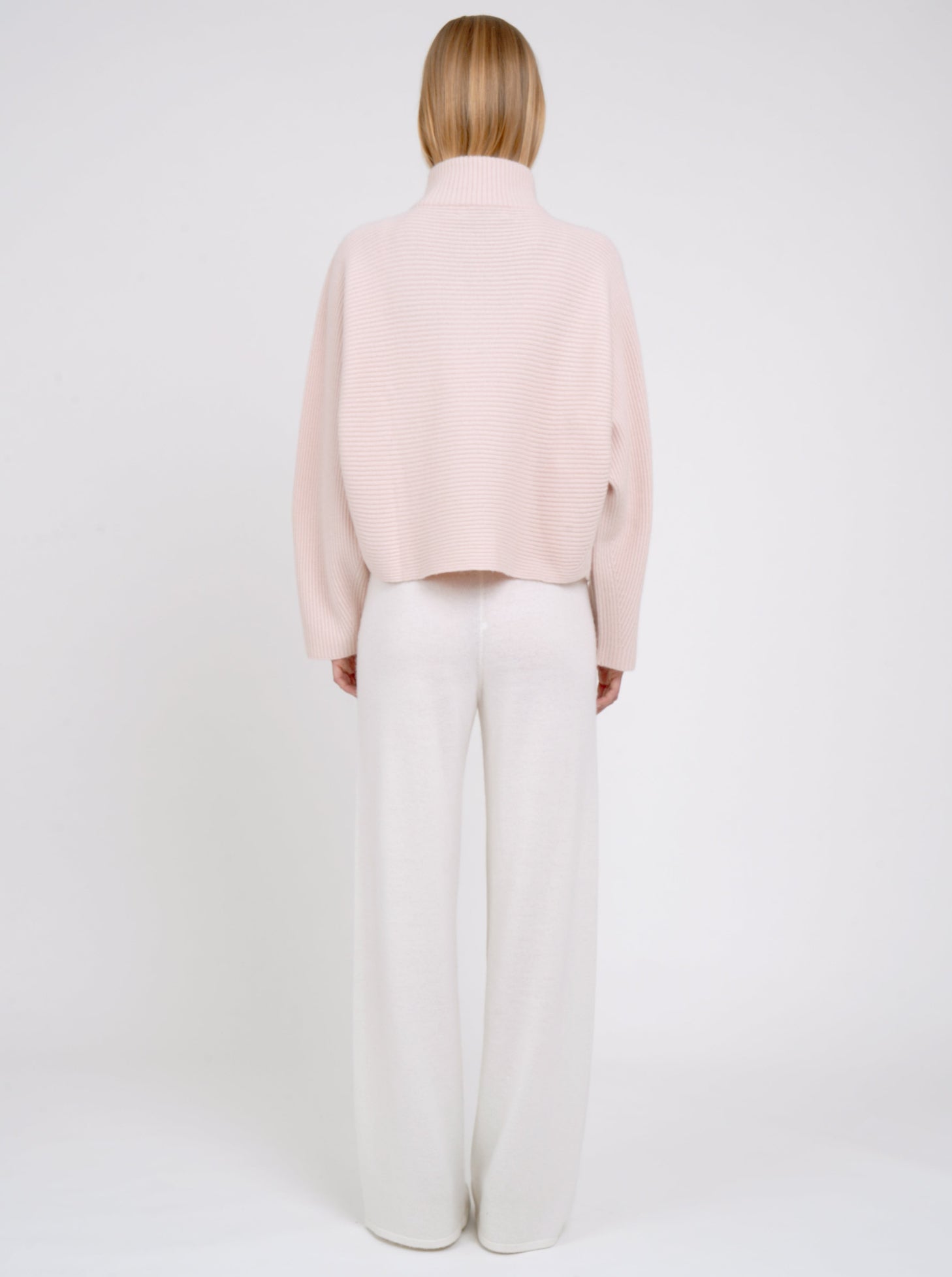 Back view modal in ribbed knit peach cashmere short jacket with high collar kimono sleeves and white wide leg knit pants.