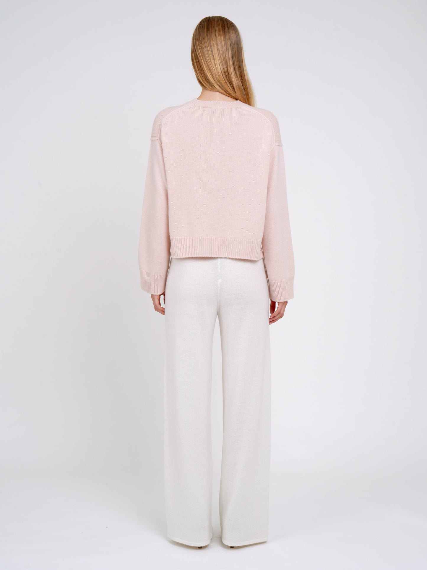 Model wearing a light pink sweater and white pants on a white background