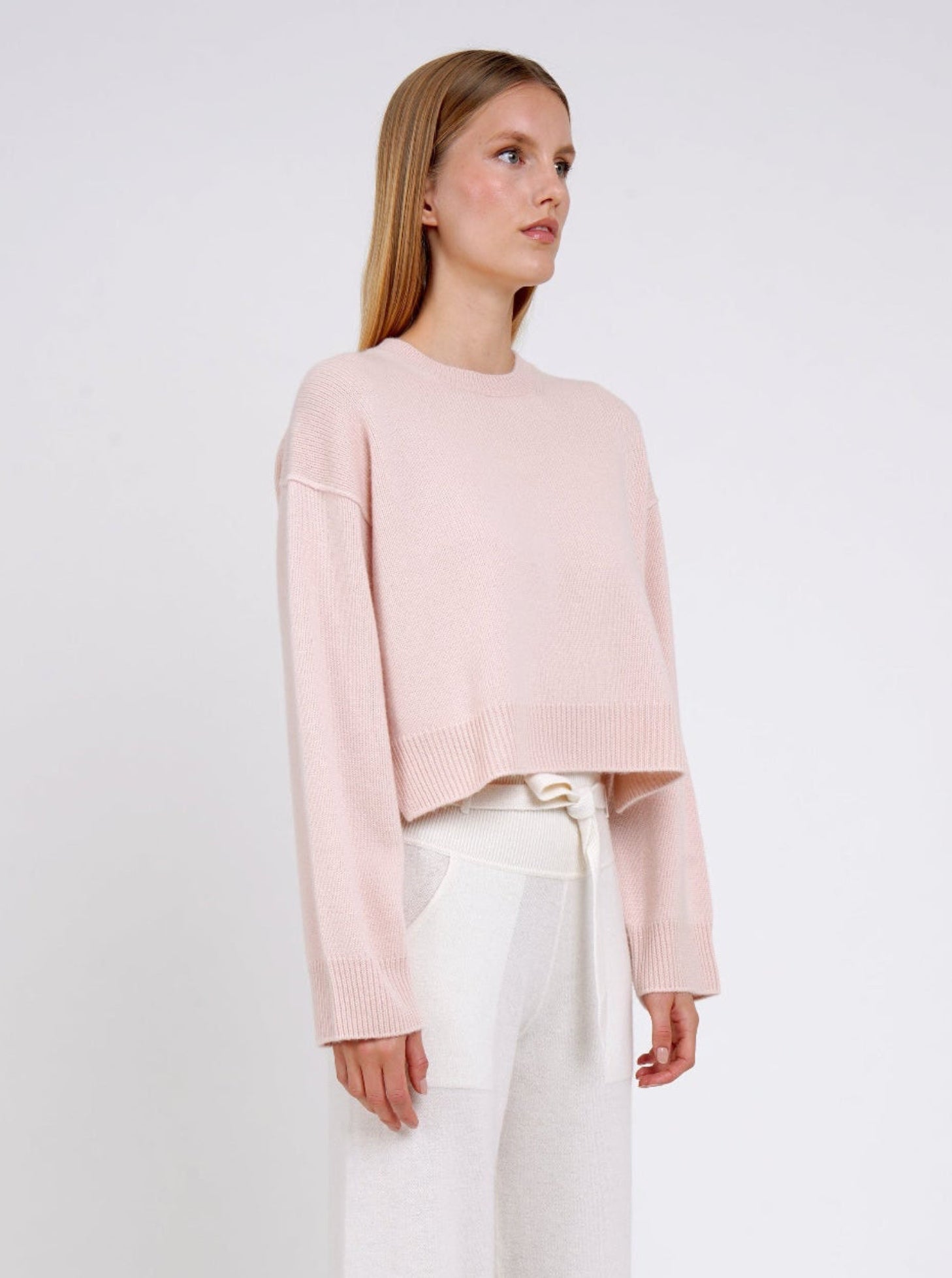 Woman wearing a light pink sweater and white pants on a white background
