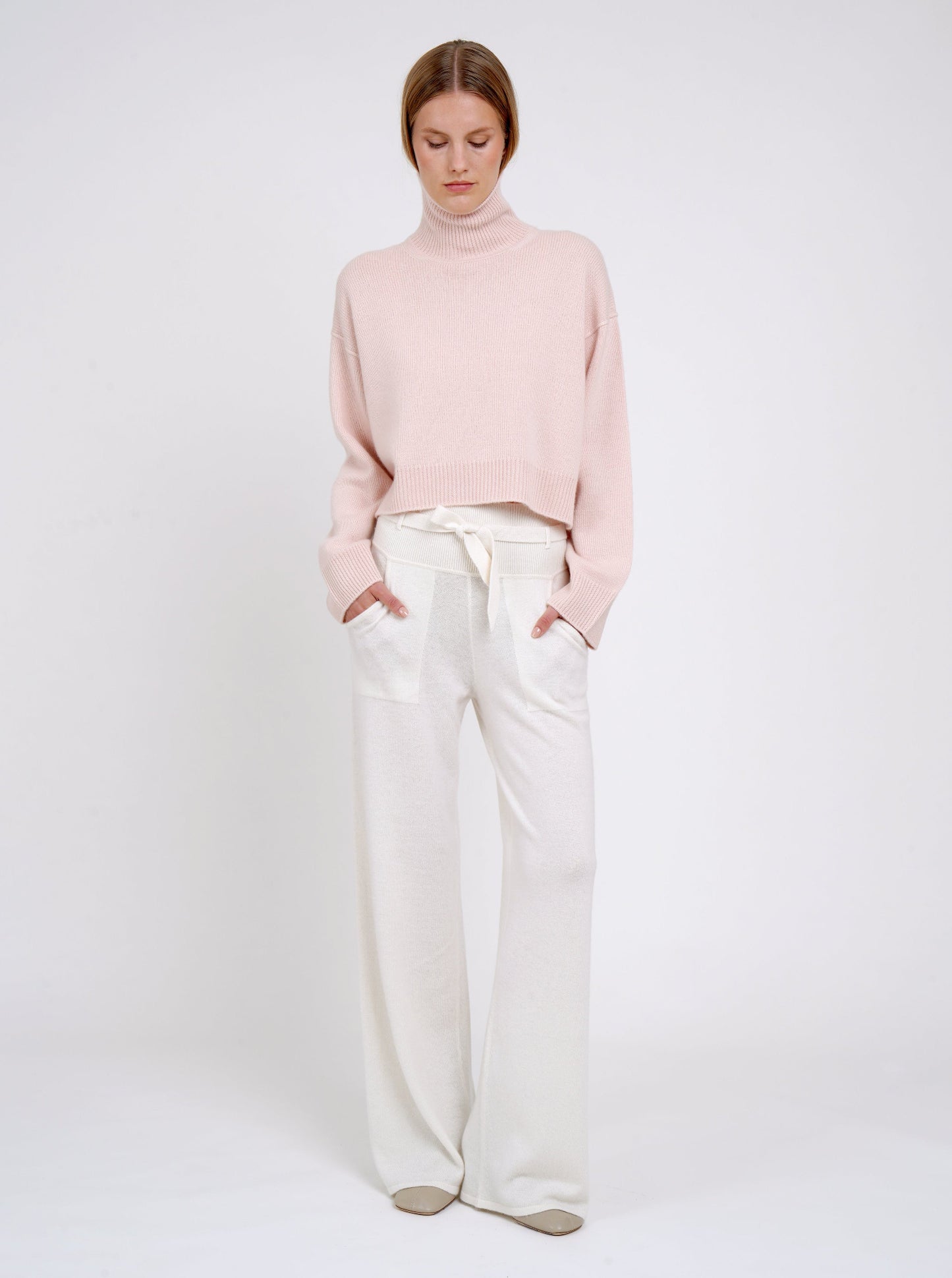 Model wearing a pink sweater and white pants on a white background