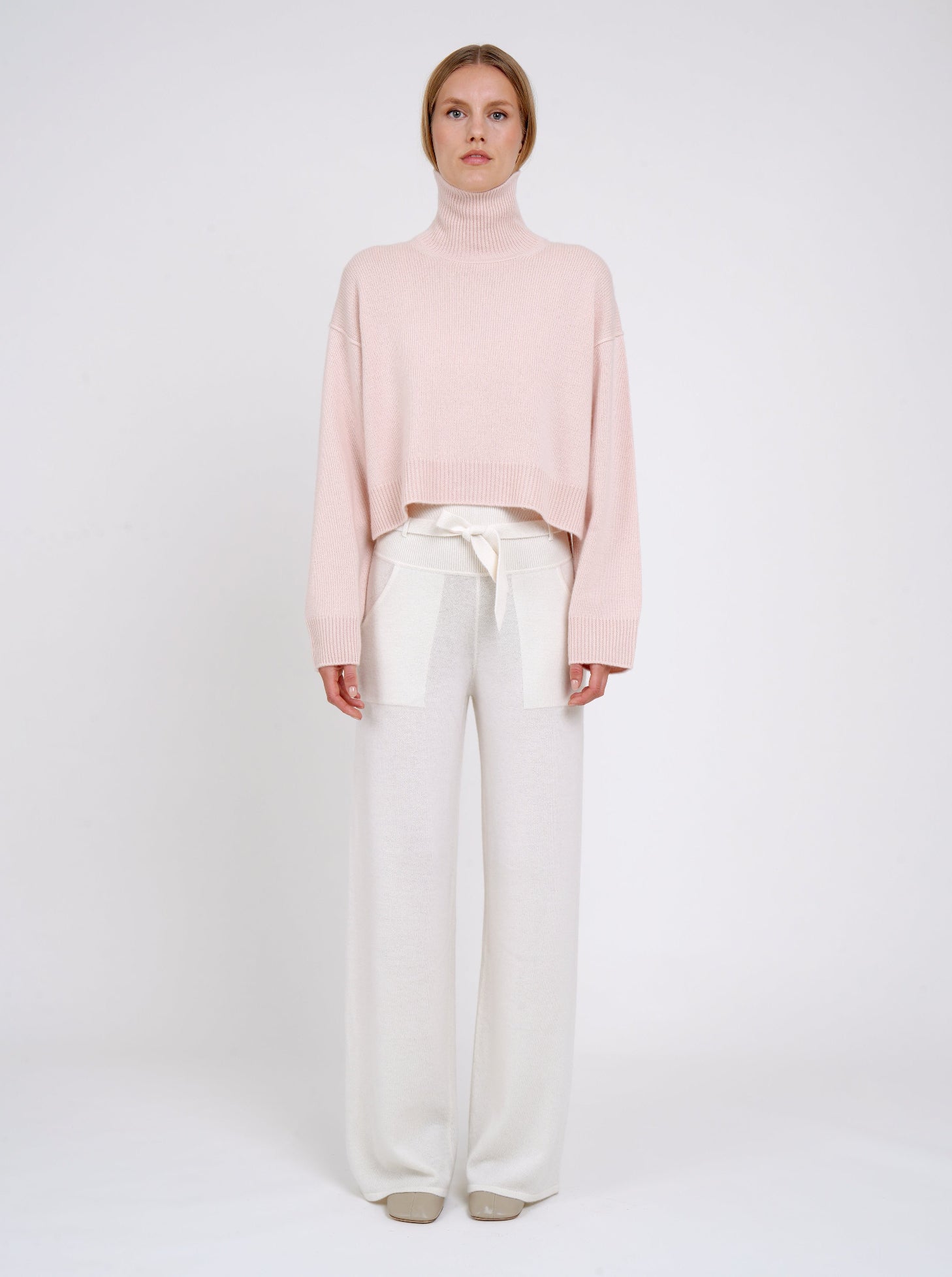 Model wearing a light pink sweater and white pants on a white background