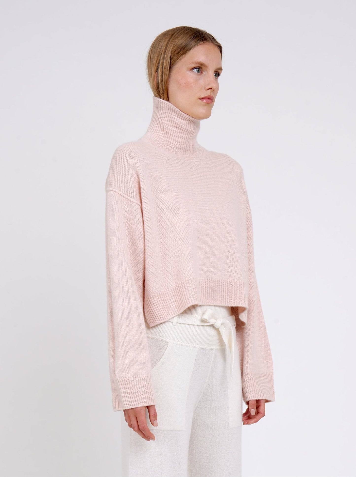 Model wearing a light pink turtleneck sweater and white pants on a plain background