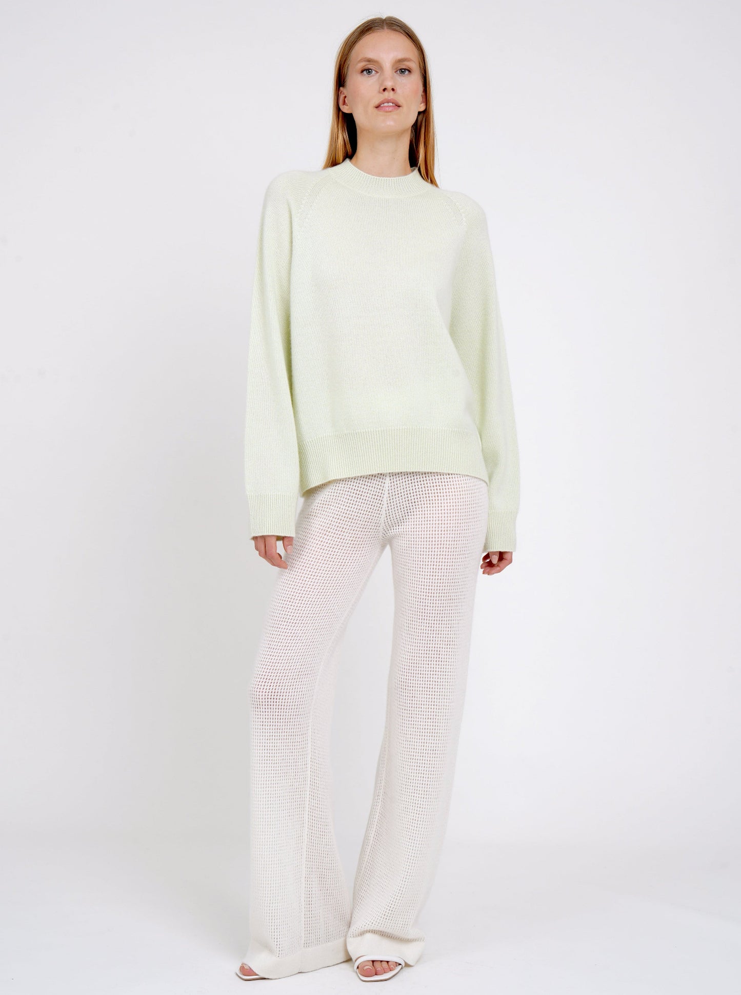Relaxed pose model in pale green cashmere jumper with round neck, long sleeves and white net knit cashmere pants.
