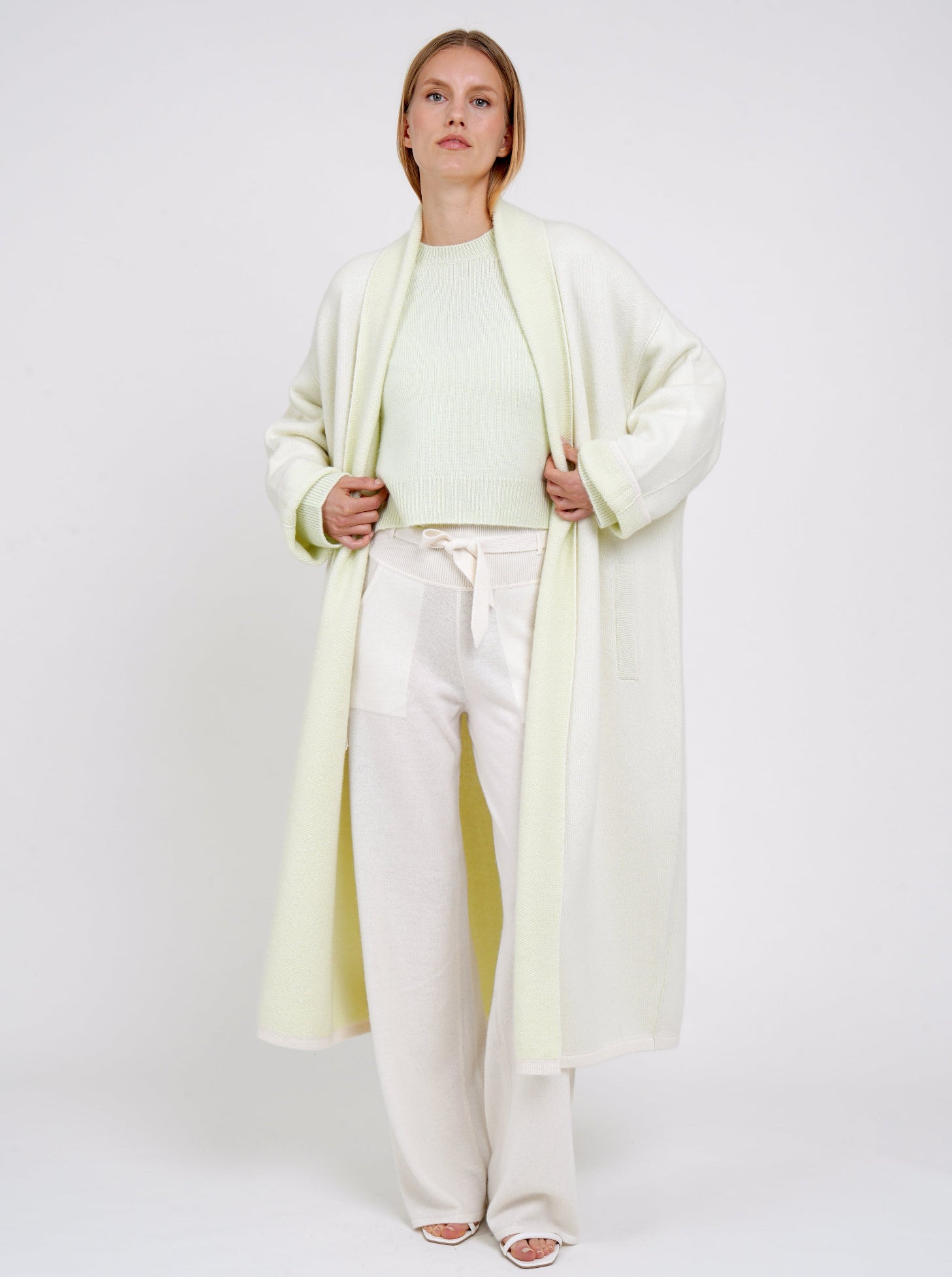 model wearing plush knit cashmere curved shape long coat in pale green with shawl collar, wide sleeves over pale green cropped jumper and white knit belted pants.