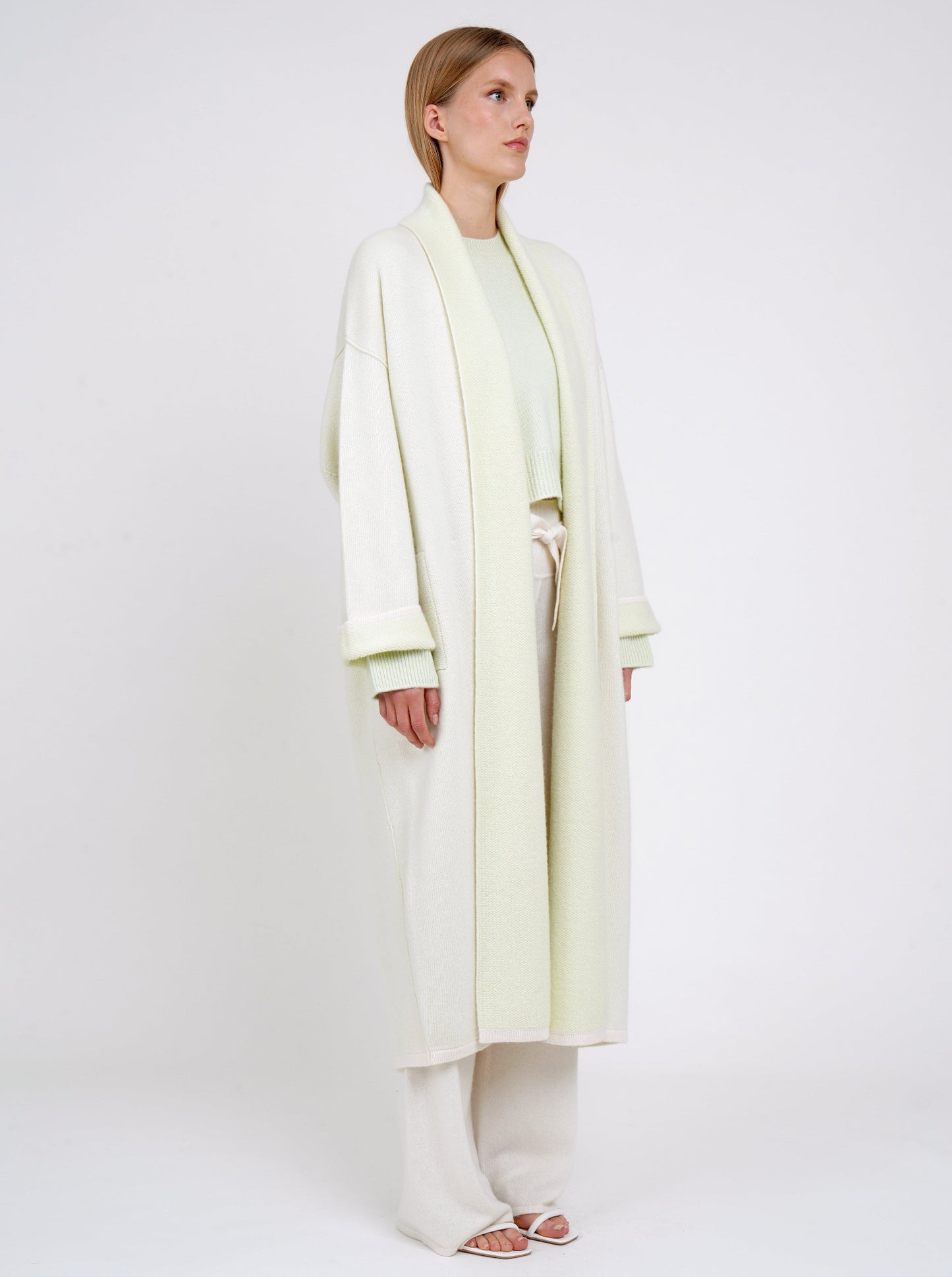 Side view model in plush knit cashmere curved shape long coat in pale green with shawl collar, wide sleeves over pale green cropped jumper and white knit pants.