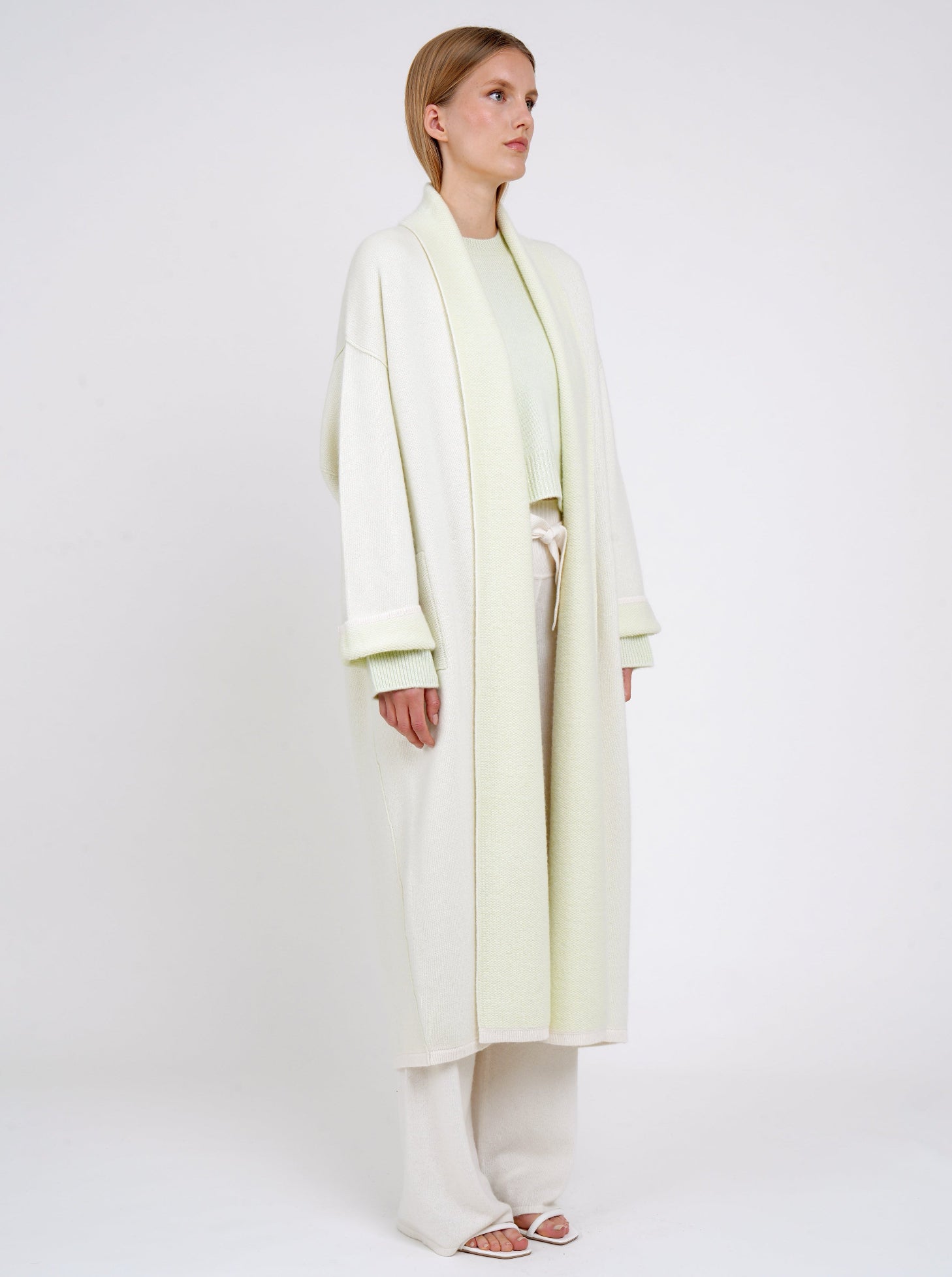 Side view model in plush knit cashmere curved shape long coat in pale green with shawl collar, wide sleeves over pale green cropped jumper and white knit pants.