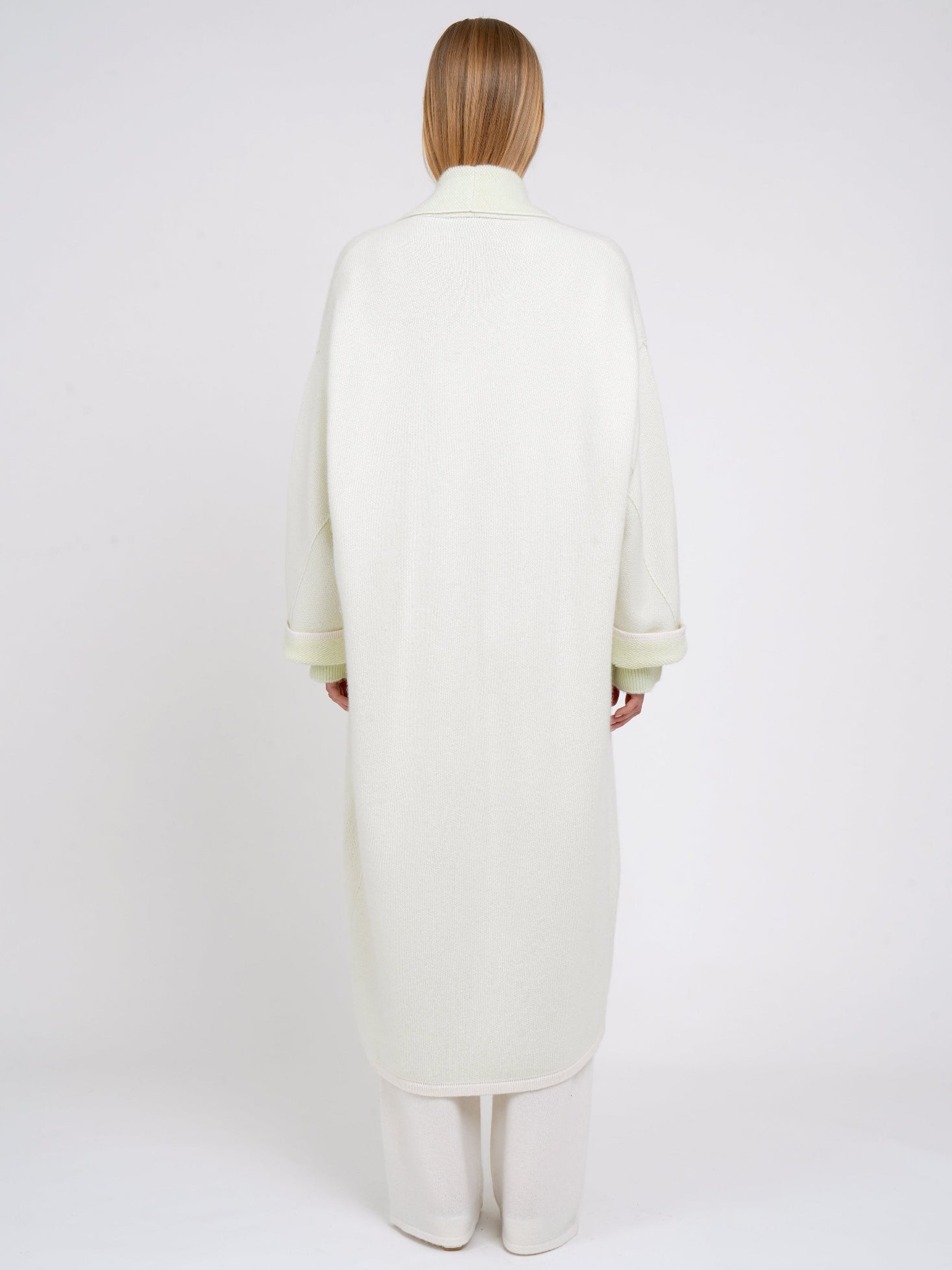back view model in plush knit pale green cashmere curved shape long coat in with shawl collar, wide sleeves over white knit pants.