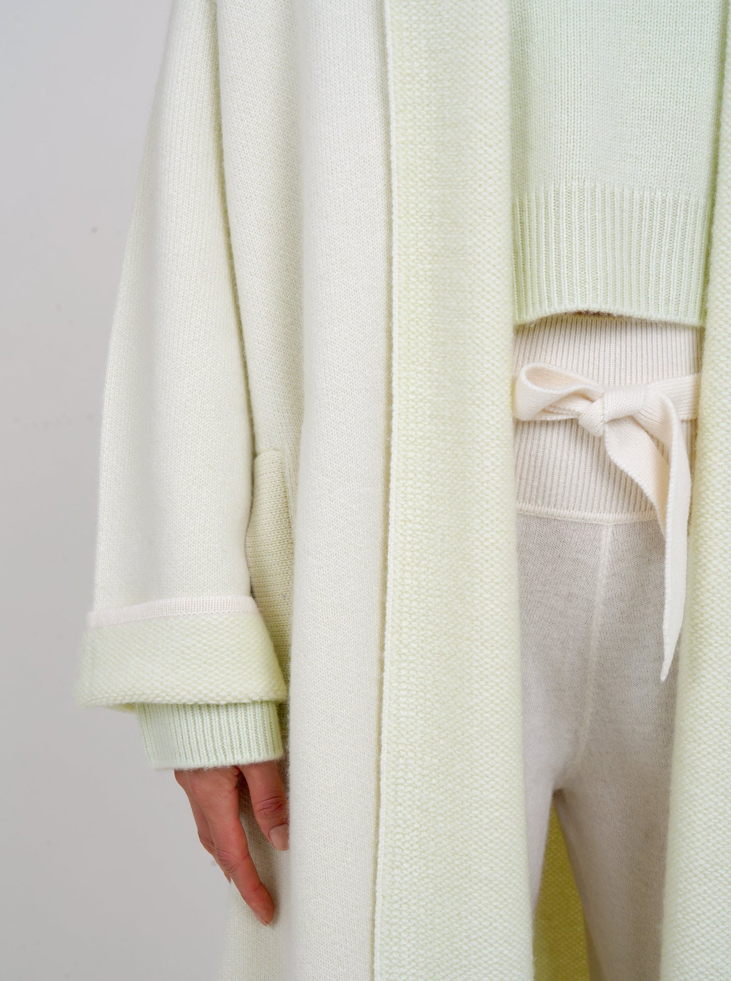 close-up detail showing plush knit cashmere coat in very pale green with shawl collar, wide sleeves over pale green cropped jumper and white knit high waist belted pants.