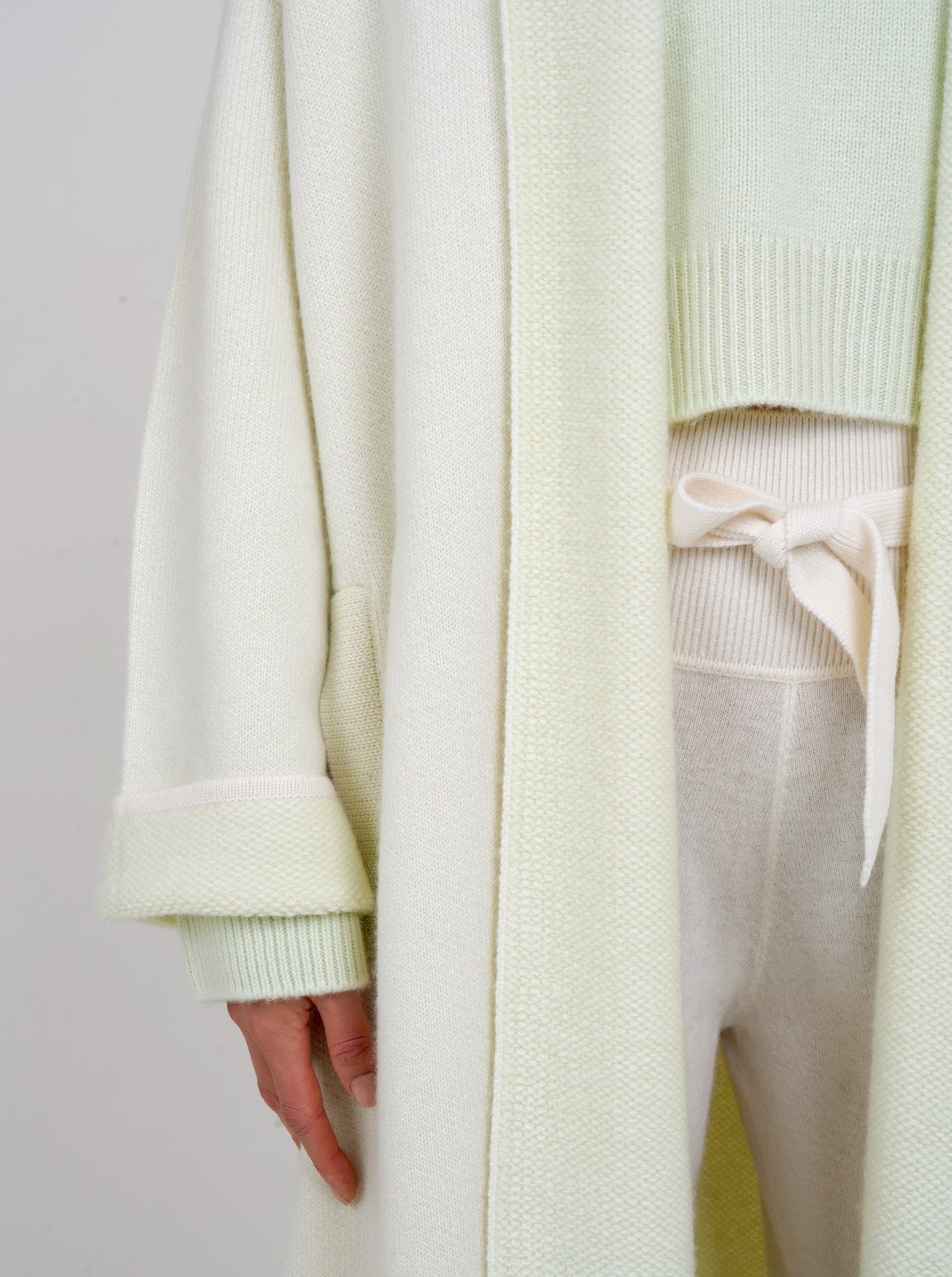 close-up detail showing plush knit cashmere coat in very pale green with shawl collar, wide sleeves over pale green cropped jumper and white knit high waist belted pants.