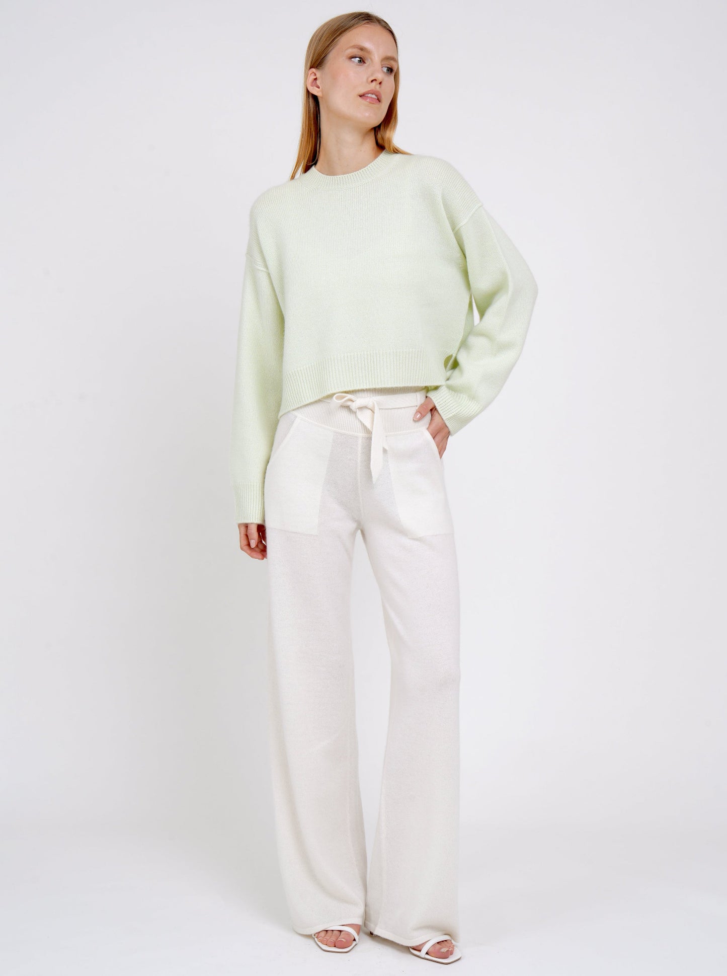Standing model posing in light green short jumper with round neck, long sleeves and white cashmere belted pants with pockets.