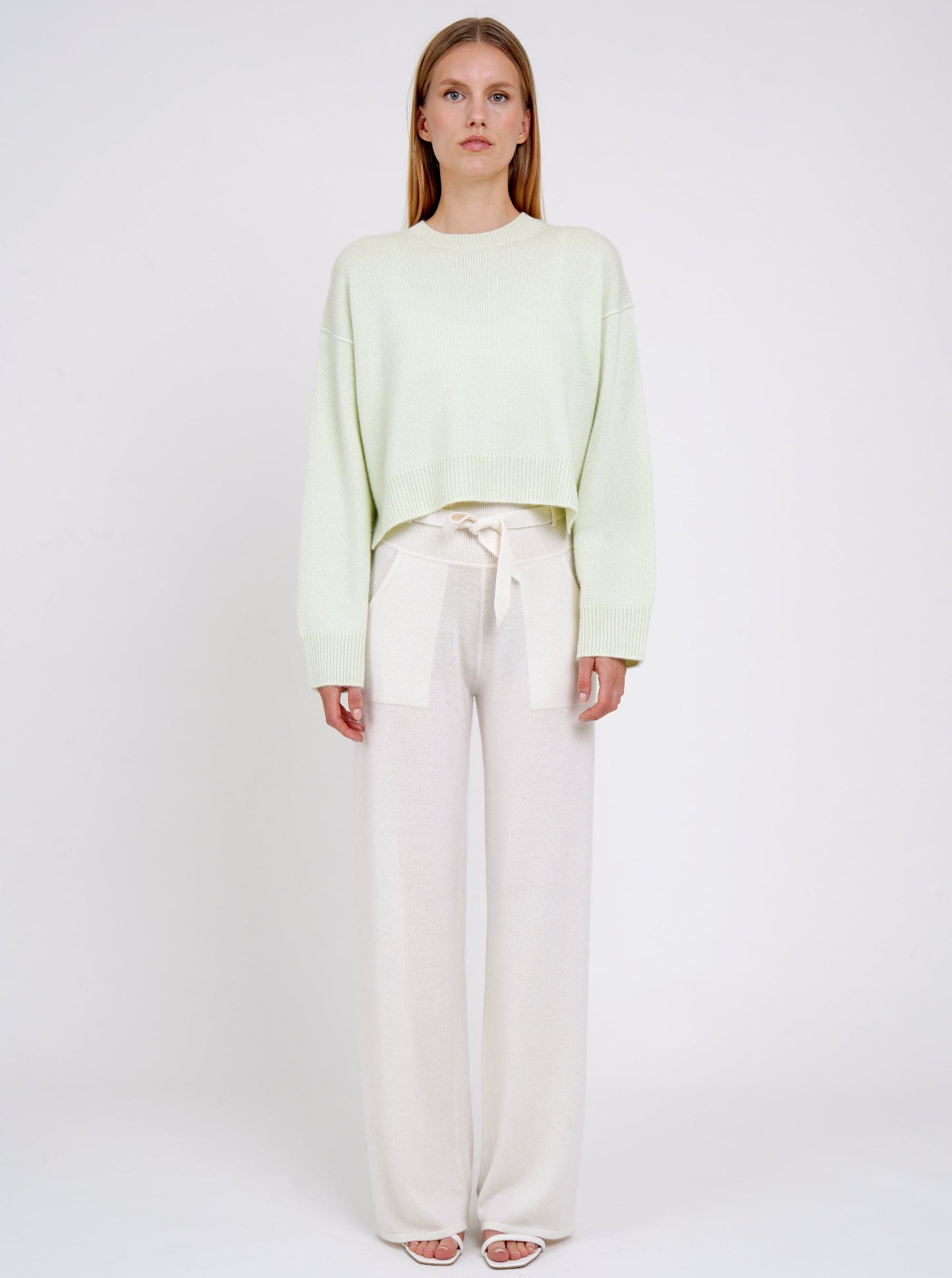 Front view model wearing light green short jumper with round neck, long sleeves and white cashmere belted wide leg pants.