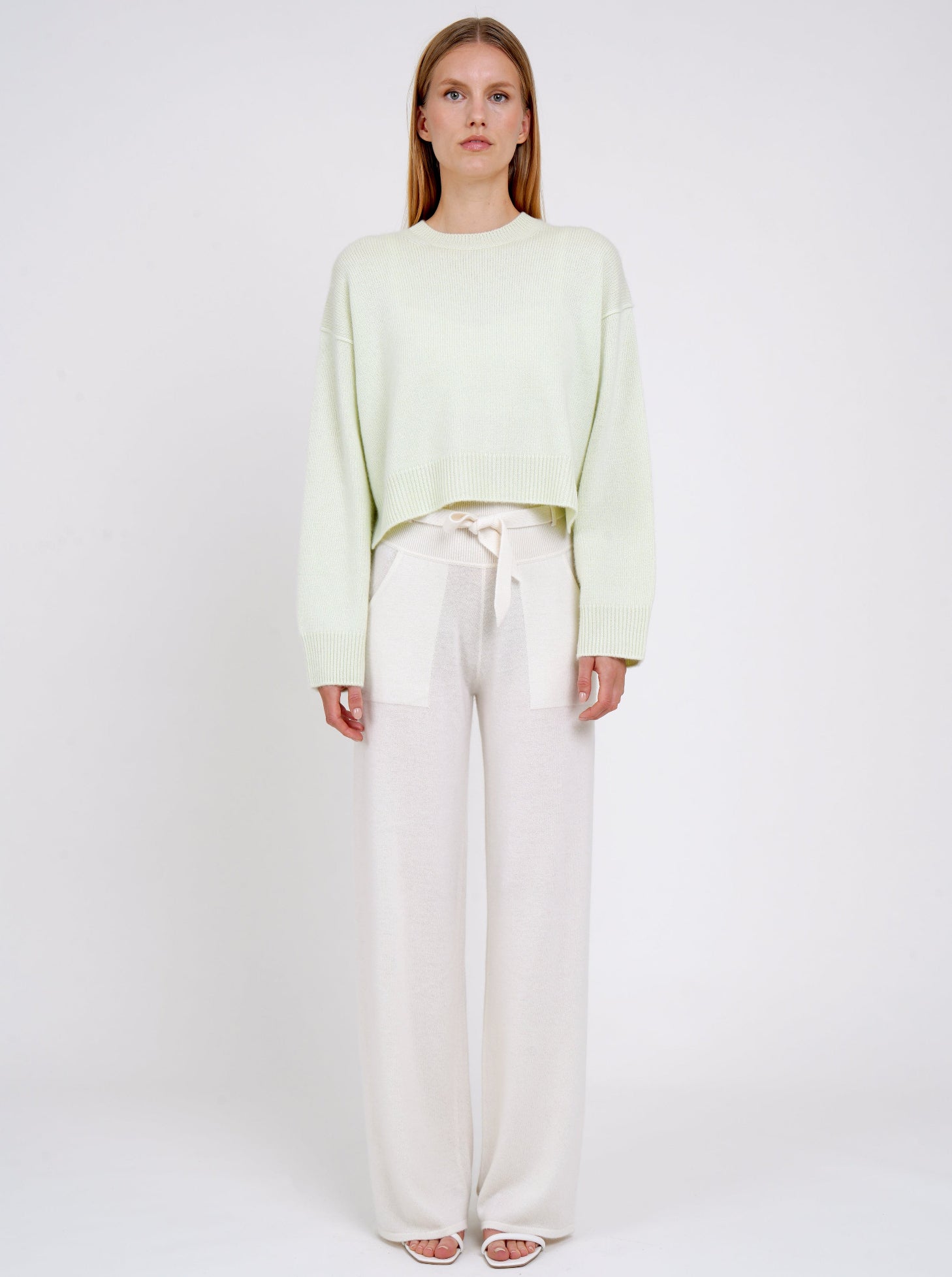 Front view model wearing light green short jumper with round neck, long sleeves and white cashmere belted wide leg pants.