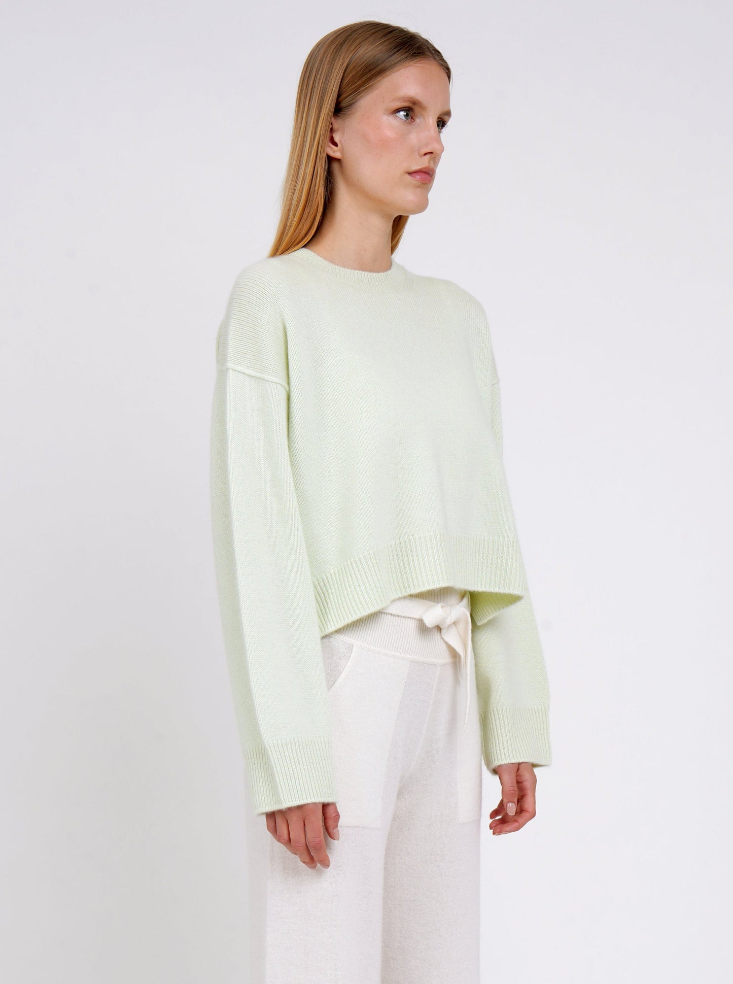Close up side view model wearing light green short jumper with round neck, long sleeves and white cashmere pants with tie belt.