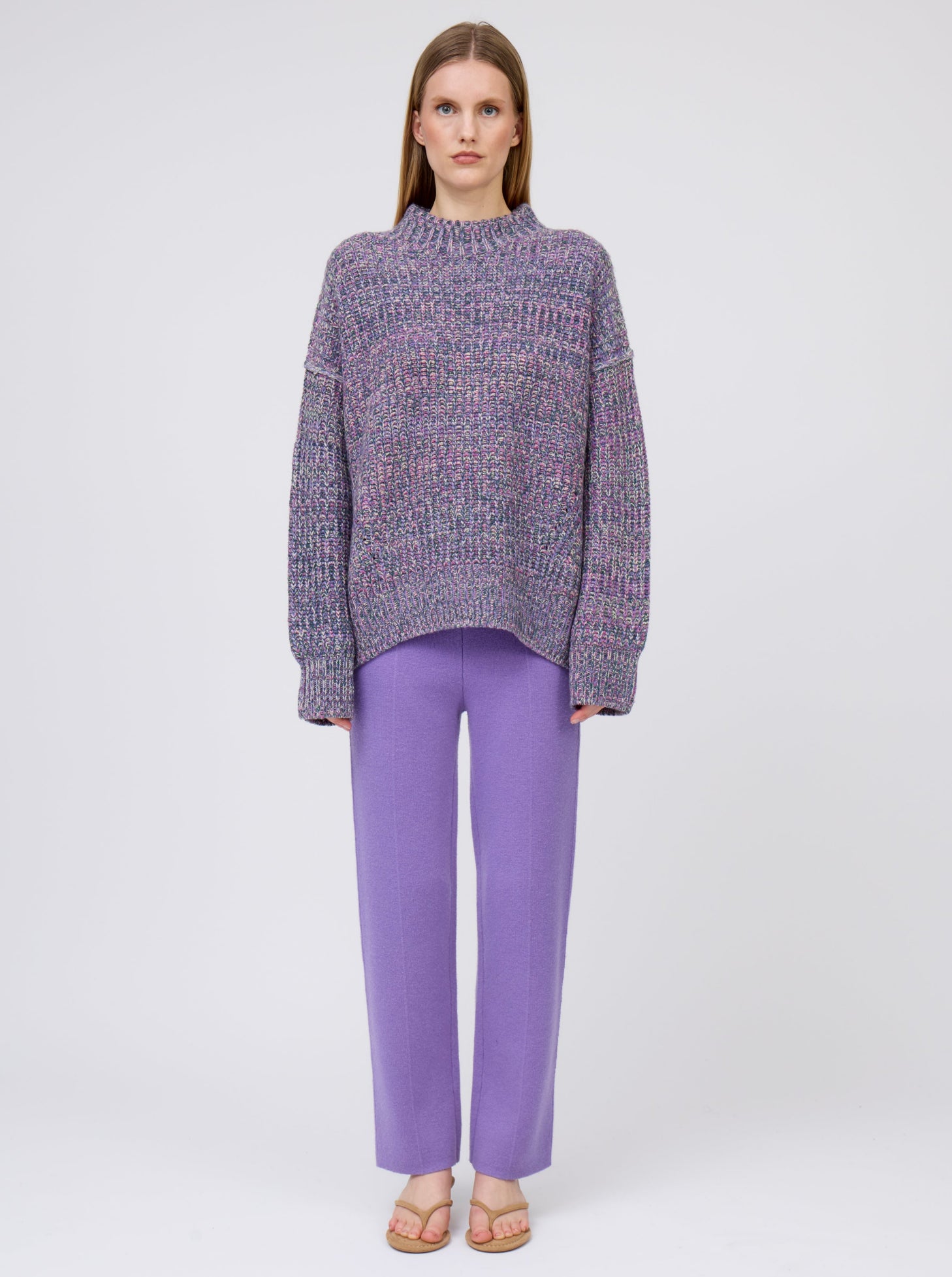 Front view model wearing chunky knit round neck sweater in mixed purple, green and yellow yarn with purple straight leg trousers.