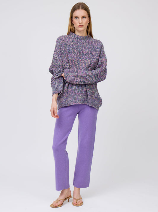 Relaxed standing pose of model wearing chunky knit round neck sweater in mixed purple, green and yellow yarn with purple straight leg trousers.