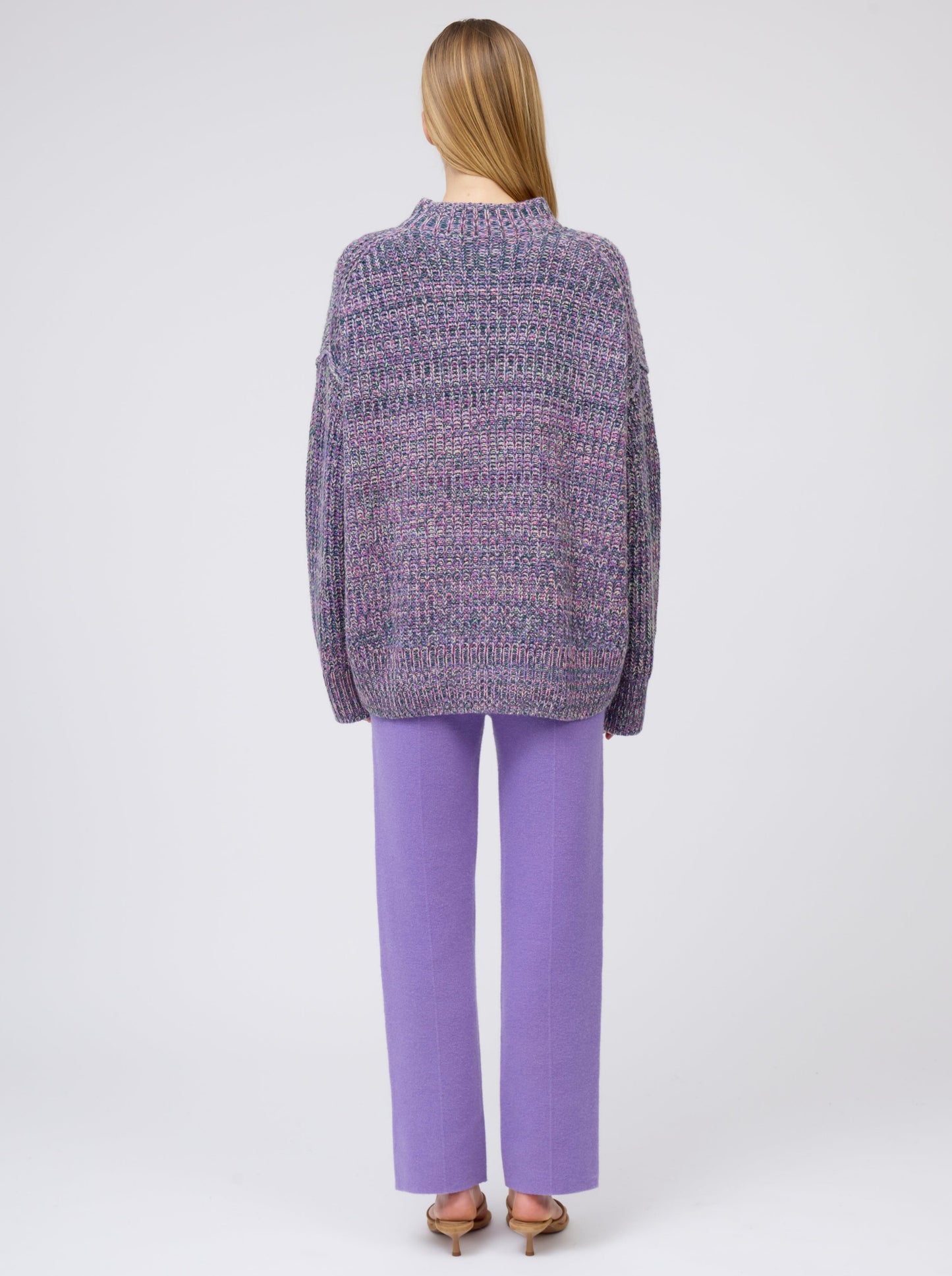 Back view model wearing chunky knit round neck sweater in mixed purple, green and yellow yarn with purple cashmere straight leg pants.