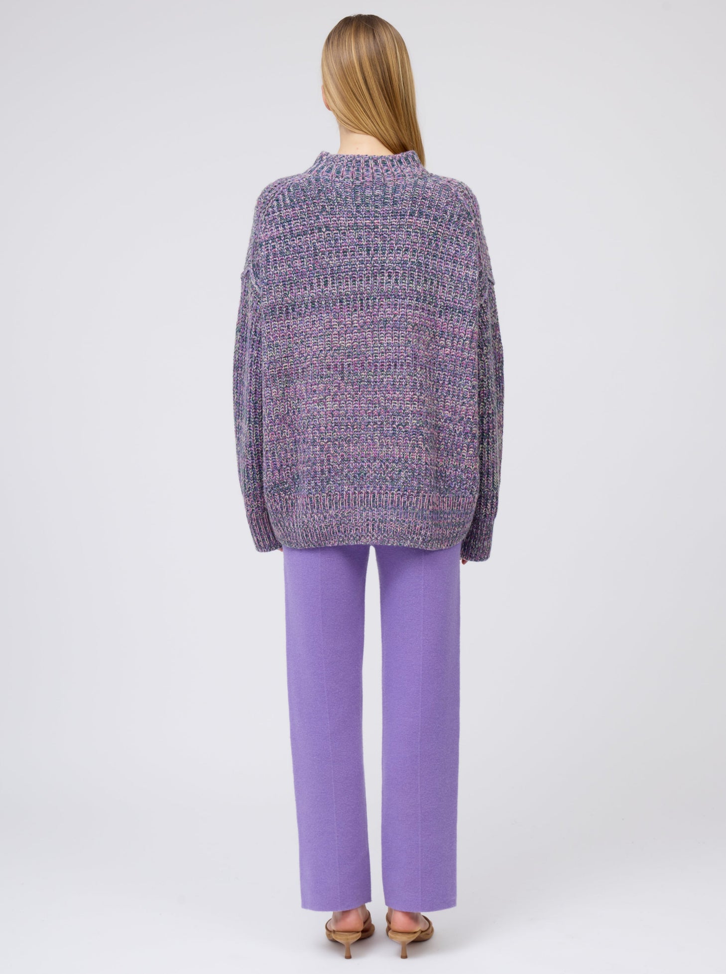 Back view model wearing chunky knit round neck sweater in mixed purple, green and yellow yarn with purple cashmere straight leg pants.