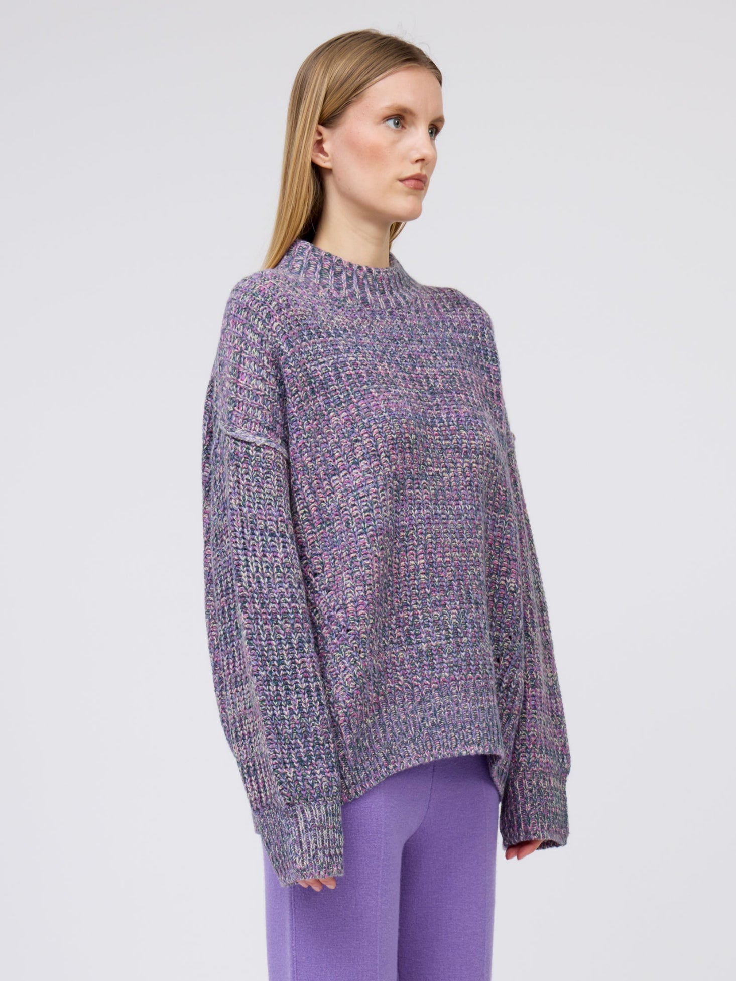 side view close up of model in chunky knit round neck sweater in mixed purple, green, pink and yellow yarn with purple cashmere straight leg pants.