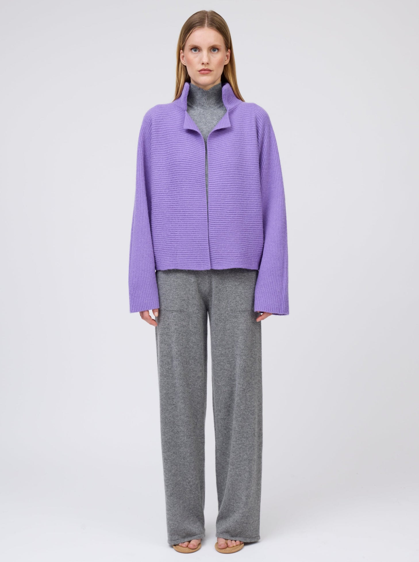 Front view model wears purple cashmere  ribbed knit jacket with kimono sleeves and high collar; worn  with grey cashmere turtleneck top and wide leg pants.