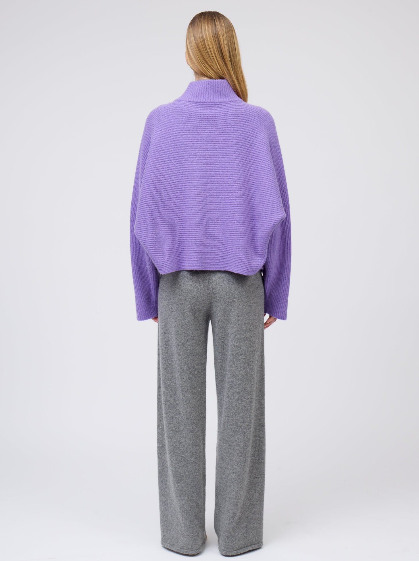 Back view model wears purple cashmere  ribbed knit jacket with kimono sleeves and high collar; worn  with grey cashmere wide leg pants.