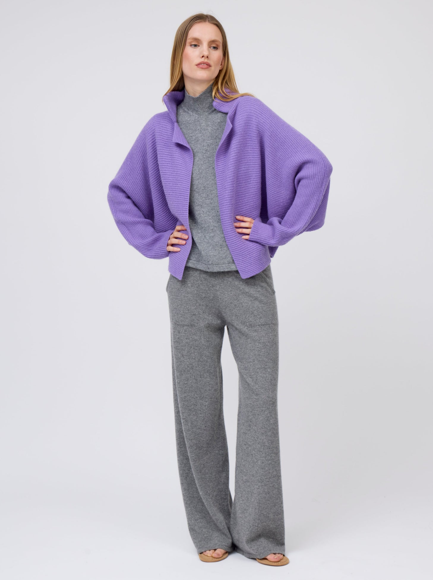 Relaxed pose model wears purple cashmere  ribbed knit jacket with kimono sleeves and high collar; worn  with grey cashmere turtleneck top and wide leg pants.