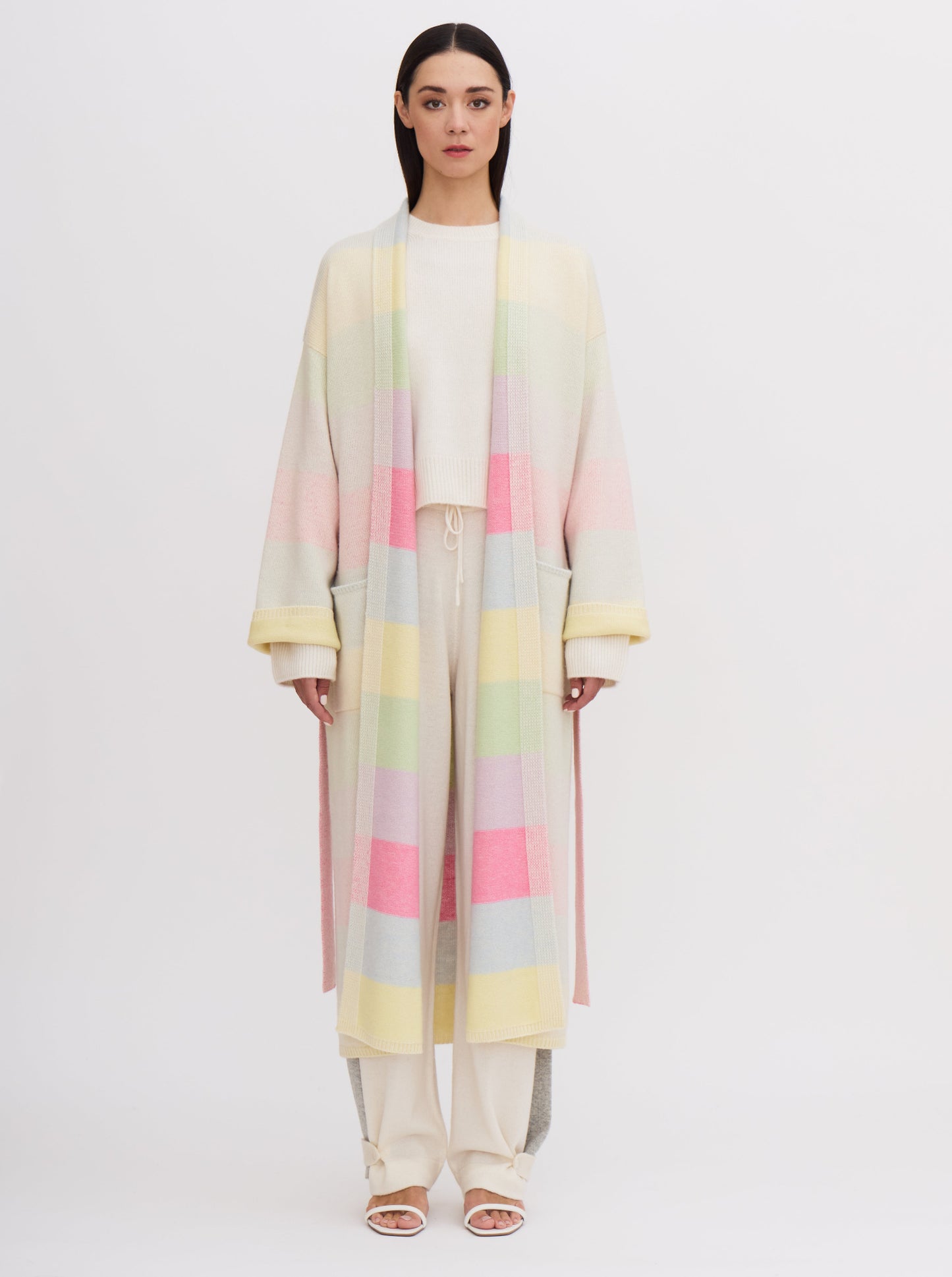 Front view model wears knitted cashmere robe coat in pastel colour rainbow stripes, over white cashmere top and pants.