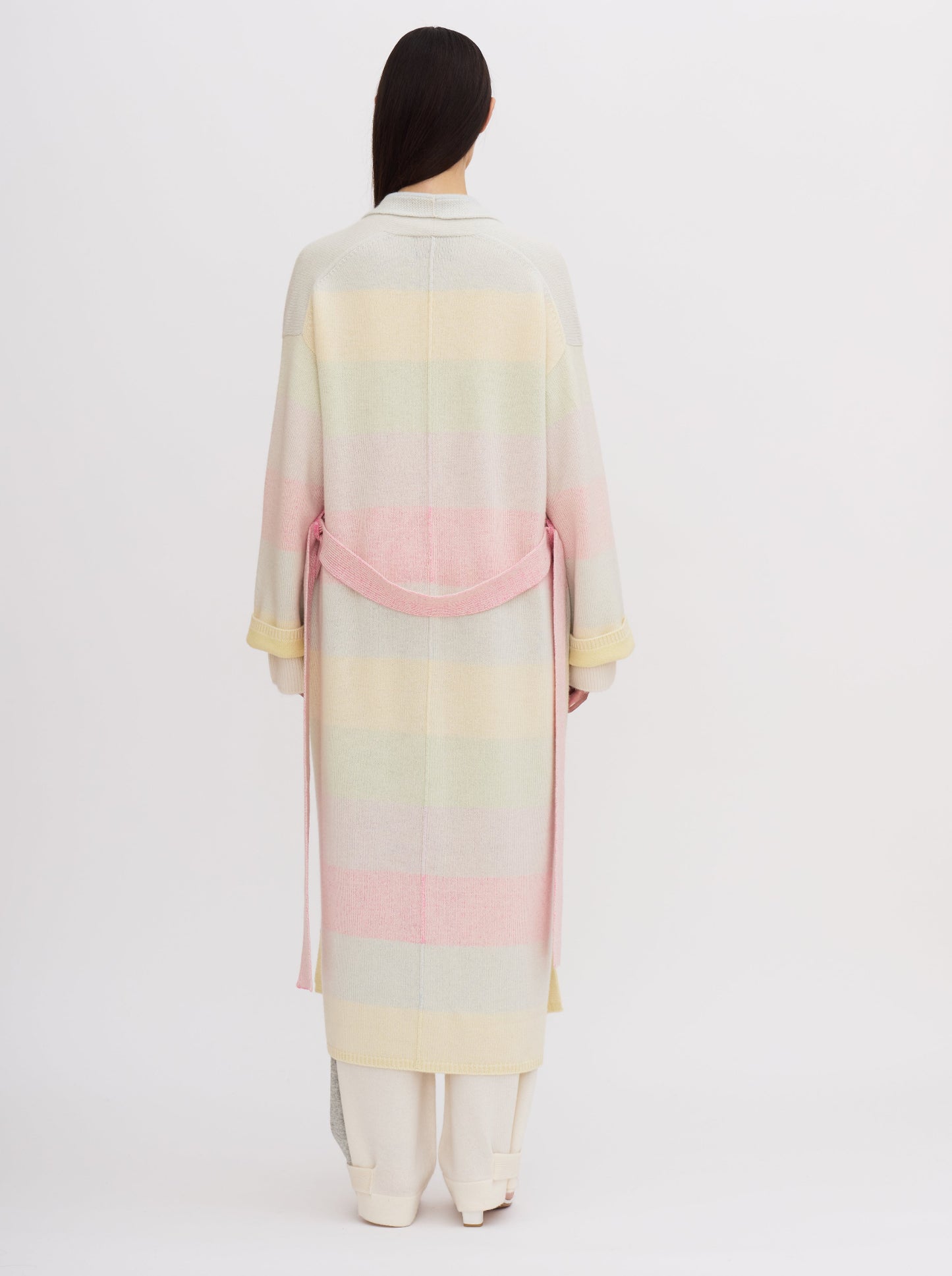 Back view model wears knitted cashmere robe coat in pastel colour rainbow stripes, with matching belt, over white cashmere top and pants.