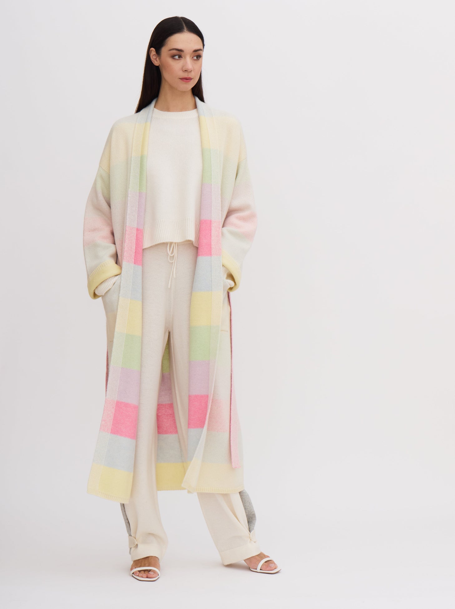 Model in relaxed pose, with hands in pockets, wears knitted cashmere robe coat in pastel colour rainbow stripes, over white cashmere top and pants.