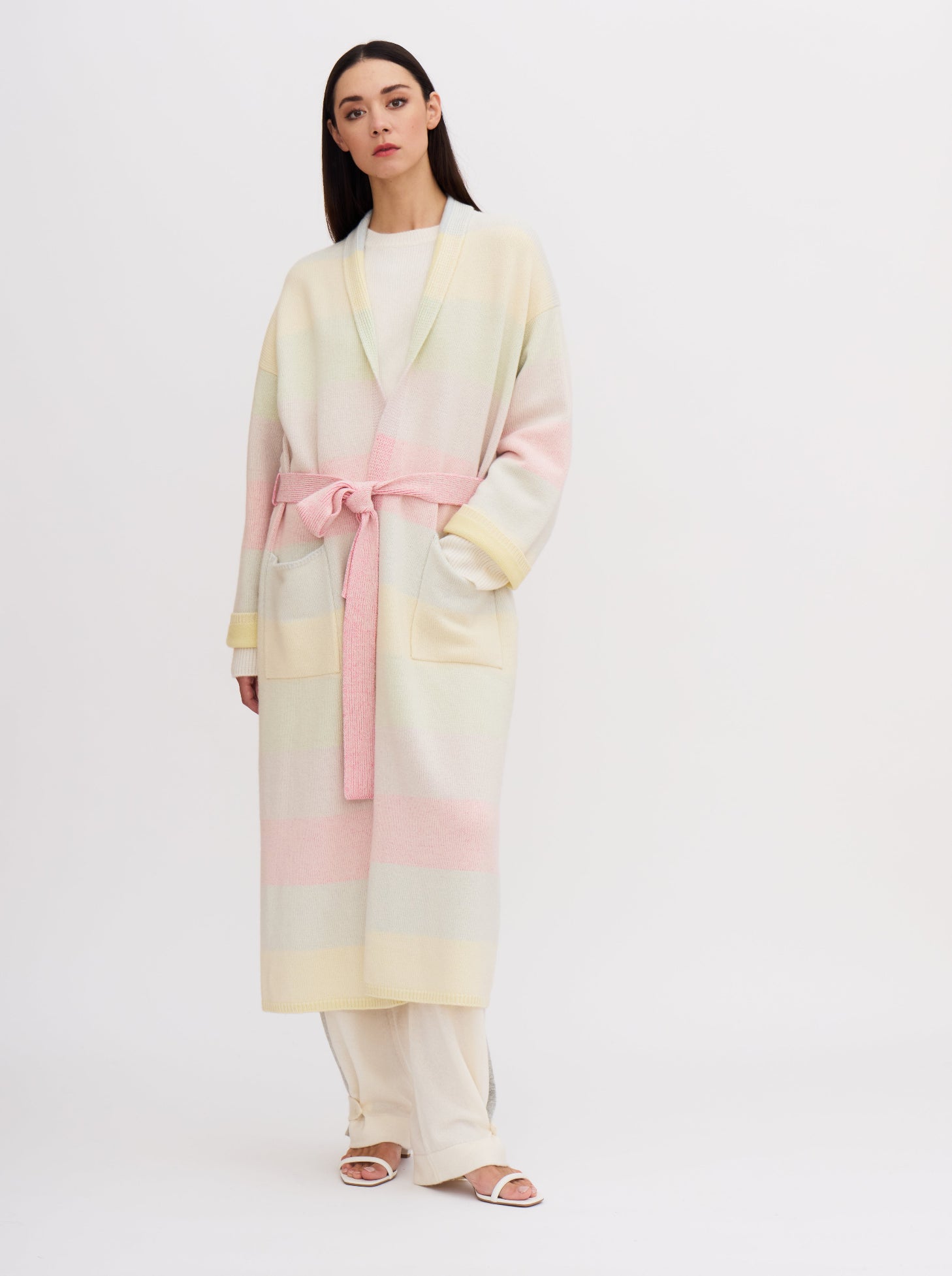 Model wears knitted cashmere robe coat in pastel colour rainbow stripes, fastened with a matching tied belt, over white cashmere knit top and pants.