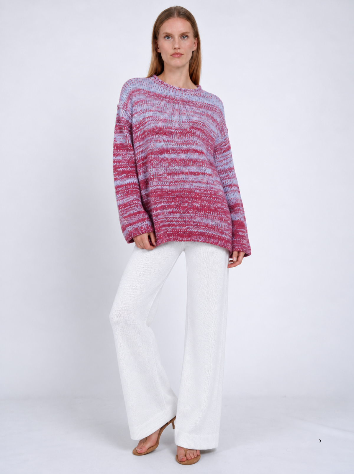 Model wearing an oversized raspberry-pink and pale-blue marled cashmere jumper with white wide-leg trousers, standing against a light studio background.