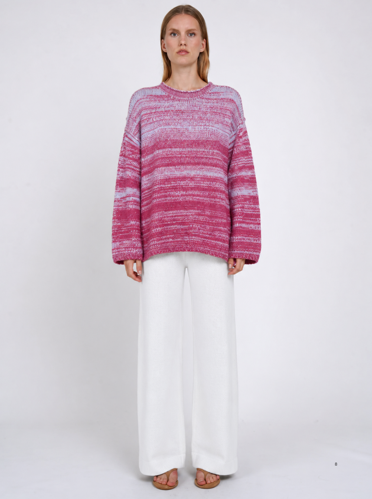 Model wearing an oversized raspberry-pink and pale-blue marled cashmere jumper with white wide-leg trousers, standing against a light studio background.