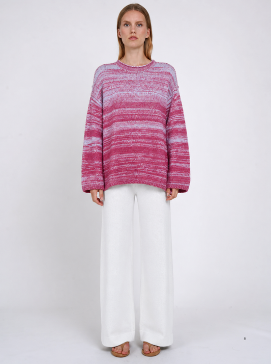Model wearing an oversized raspberry-pink and pale-blue marled cashmere jumper with white wide-leg trousers, standing against a light studio background.