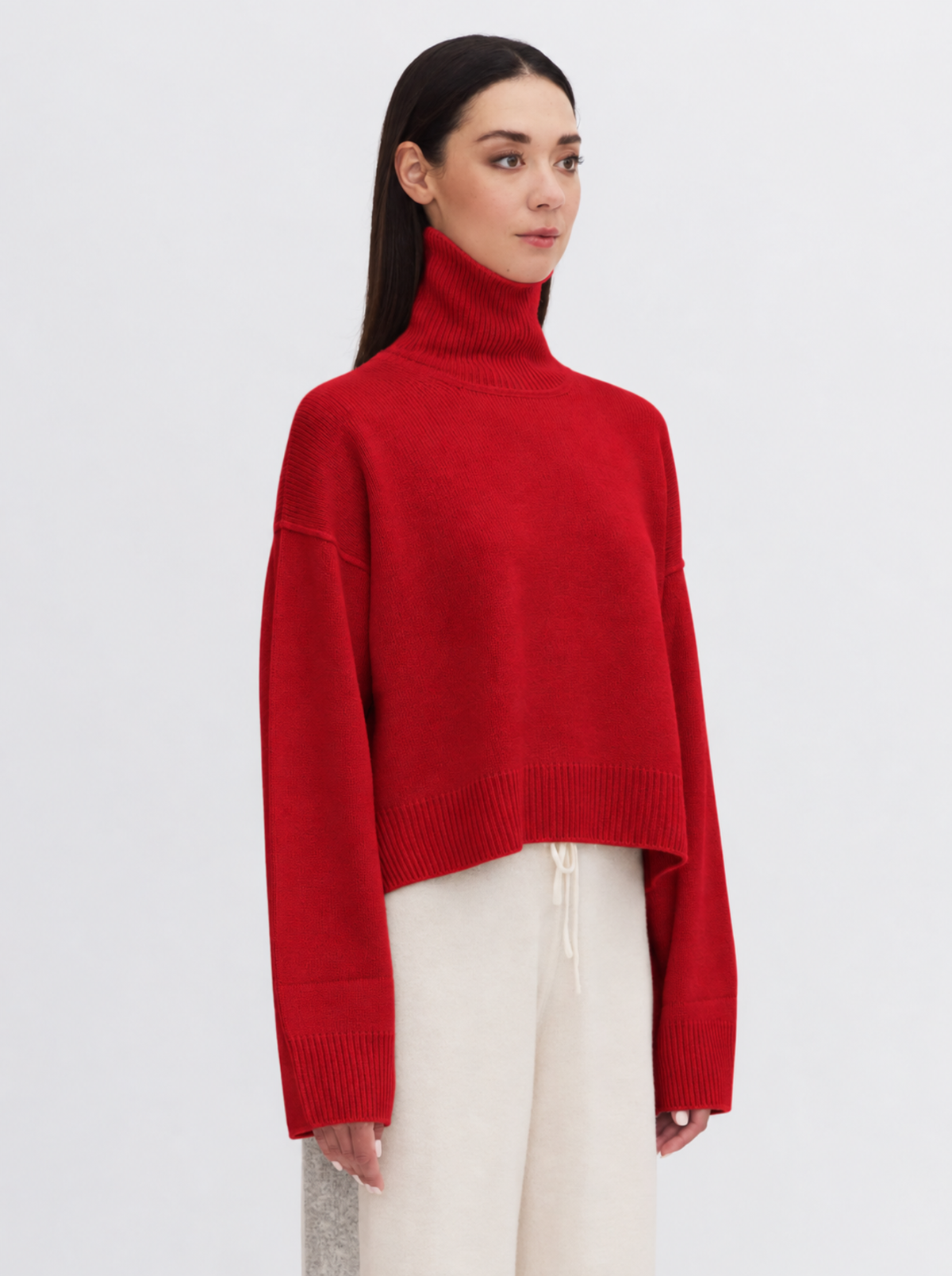 True red cropped turtleneck sweater and cream drawstring trousers, standing against a plain light gray studio background.