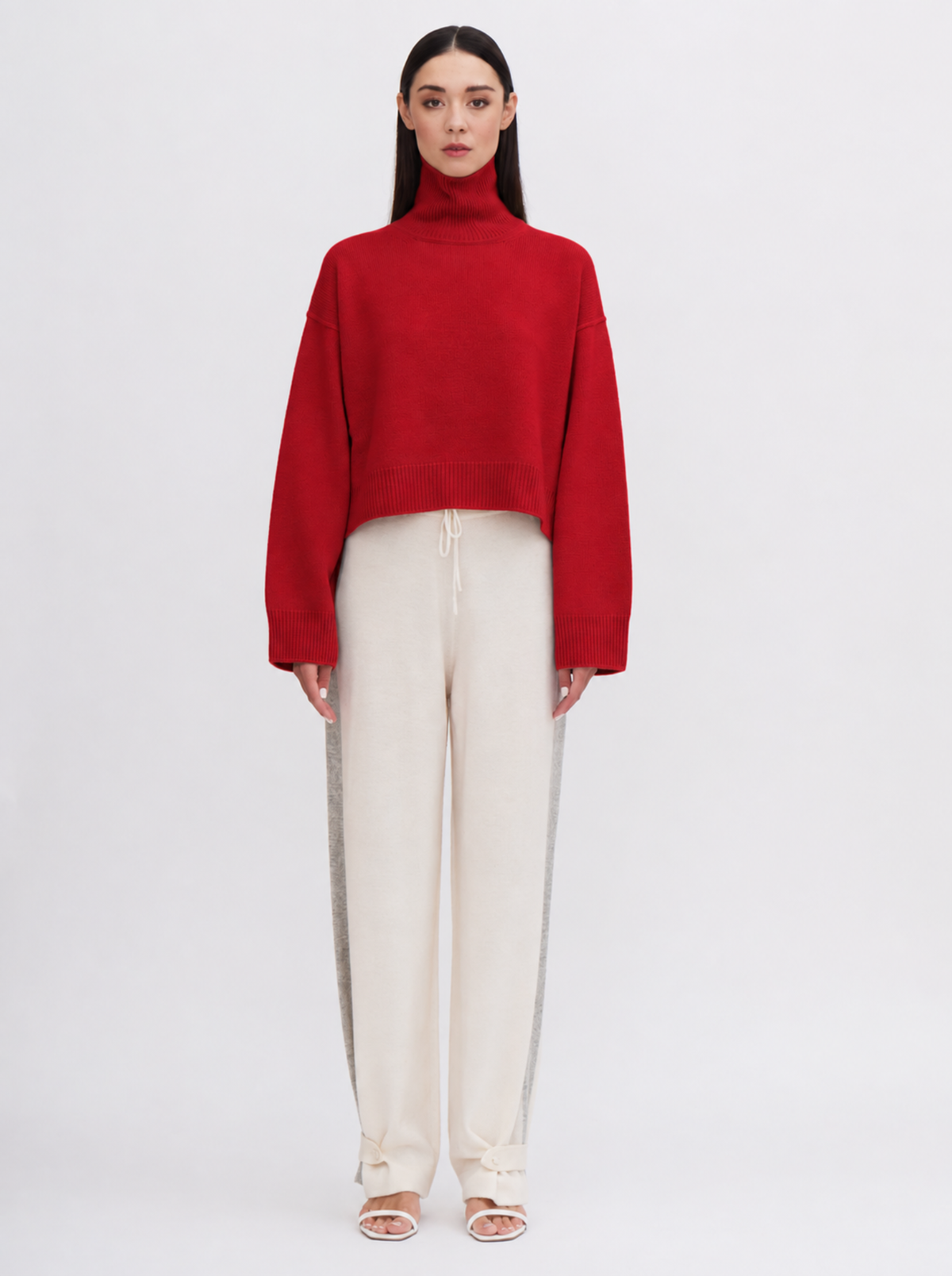 True red cropped turtleneck sweater and cream drawstring trousers, standing against a plain light gray studio background.