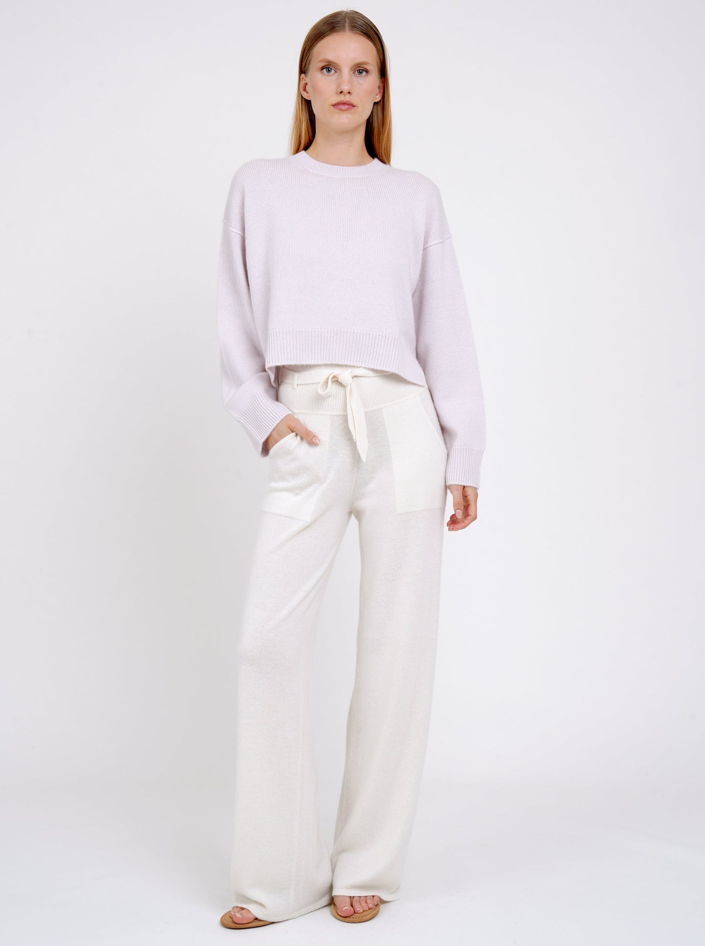 Model wears pure white cashmere, wide leg pants with knitted belt tied at the waist and lilac cashmere cropped, round neck jumper. 