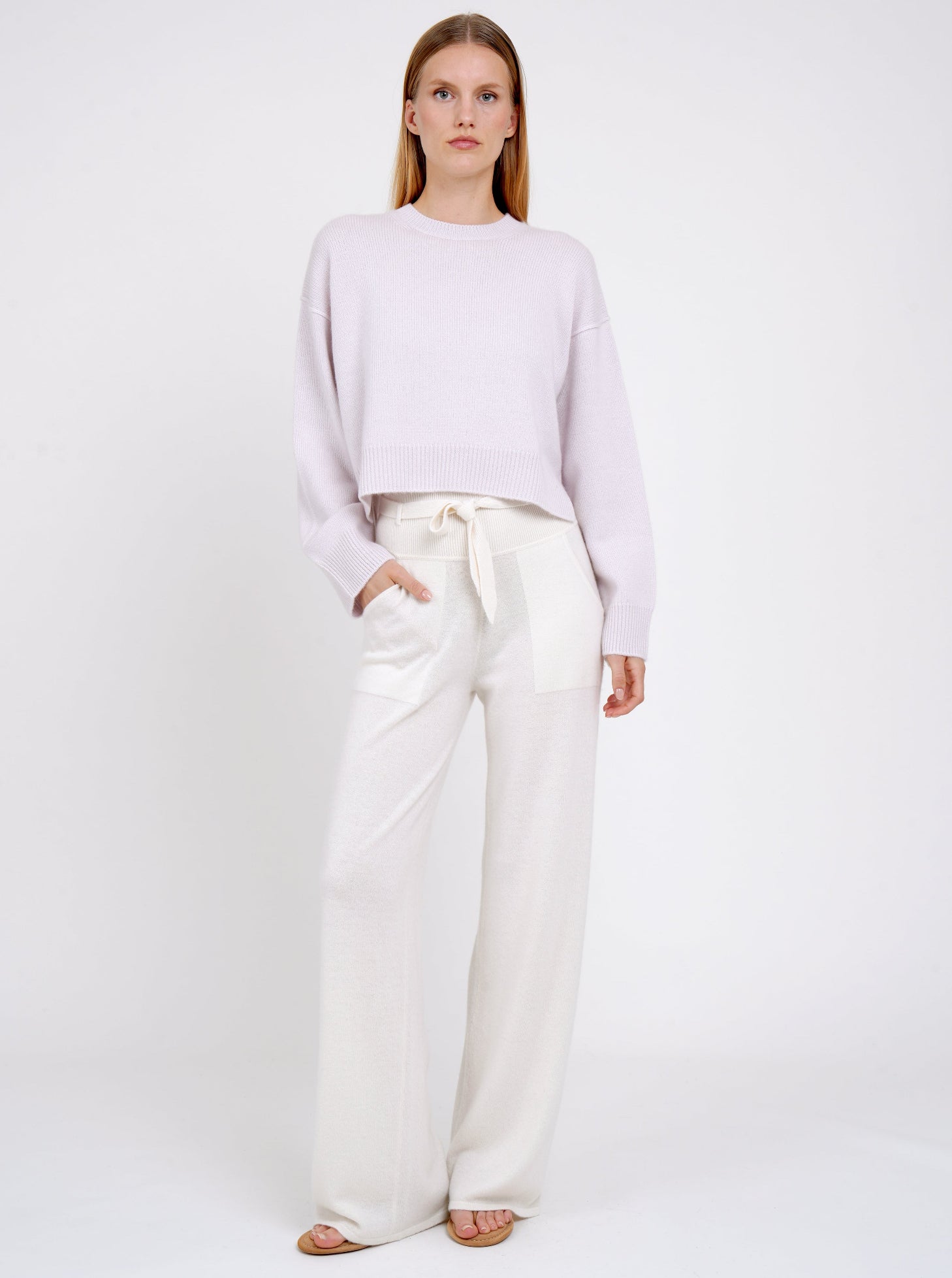 Model wears pure white cashmere, wide leg pants with knitted belt tied at the waist and lilac cashmere cropped, round neck jumper. 