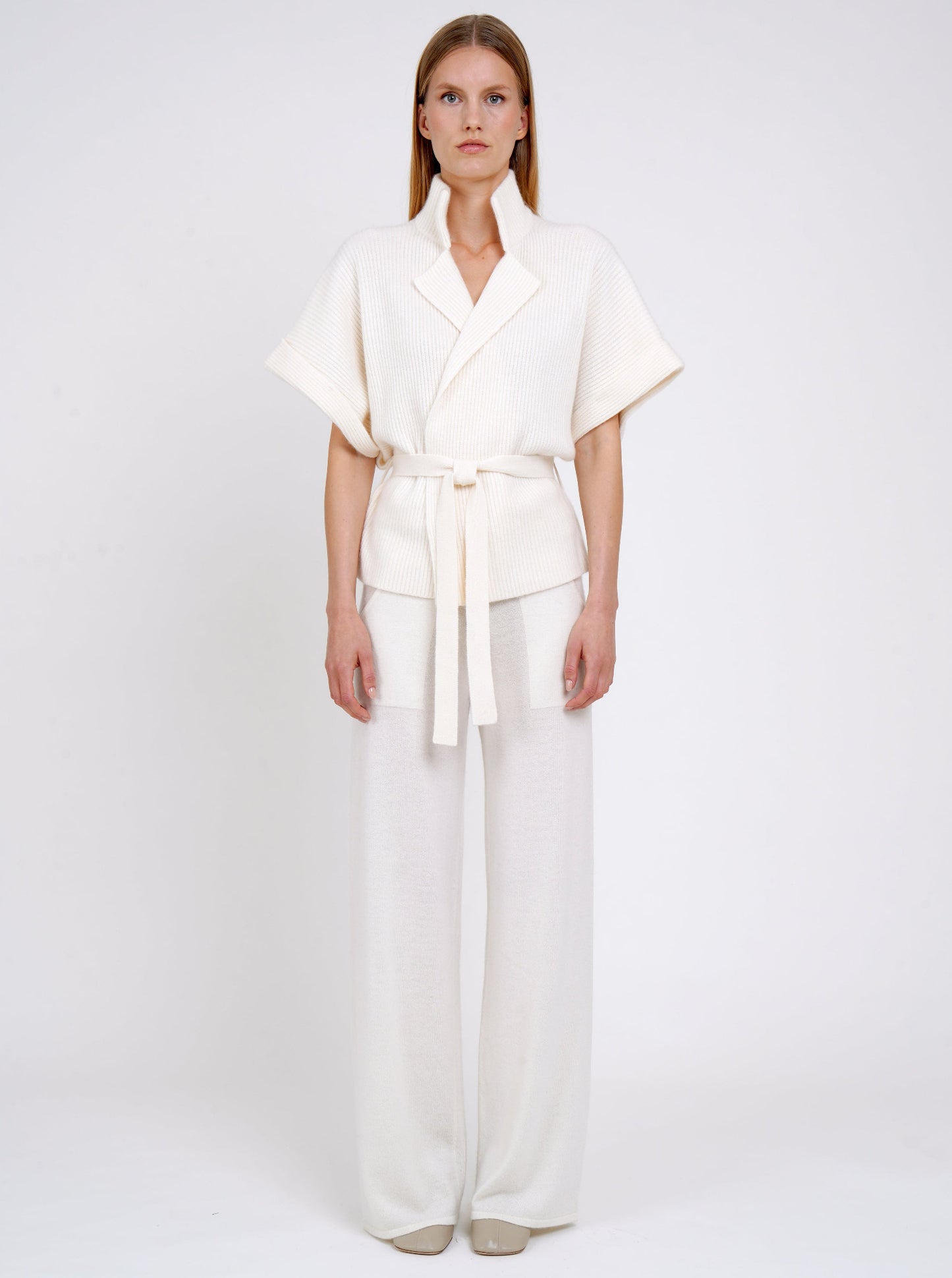 Front view model in white ribbed knit cashmere belted jacket with high collar wide short sleeves and white wide leg knit pants.