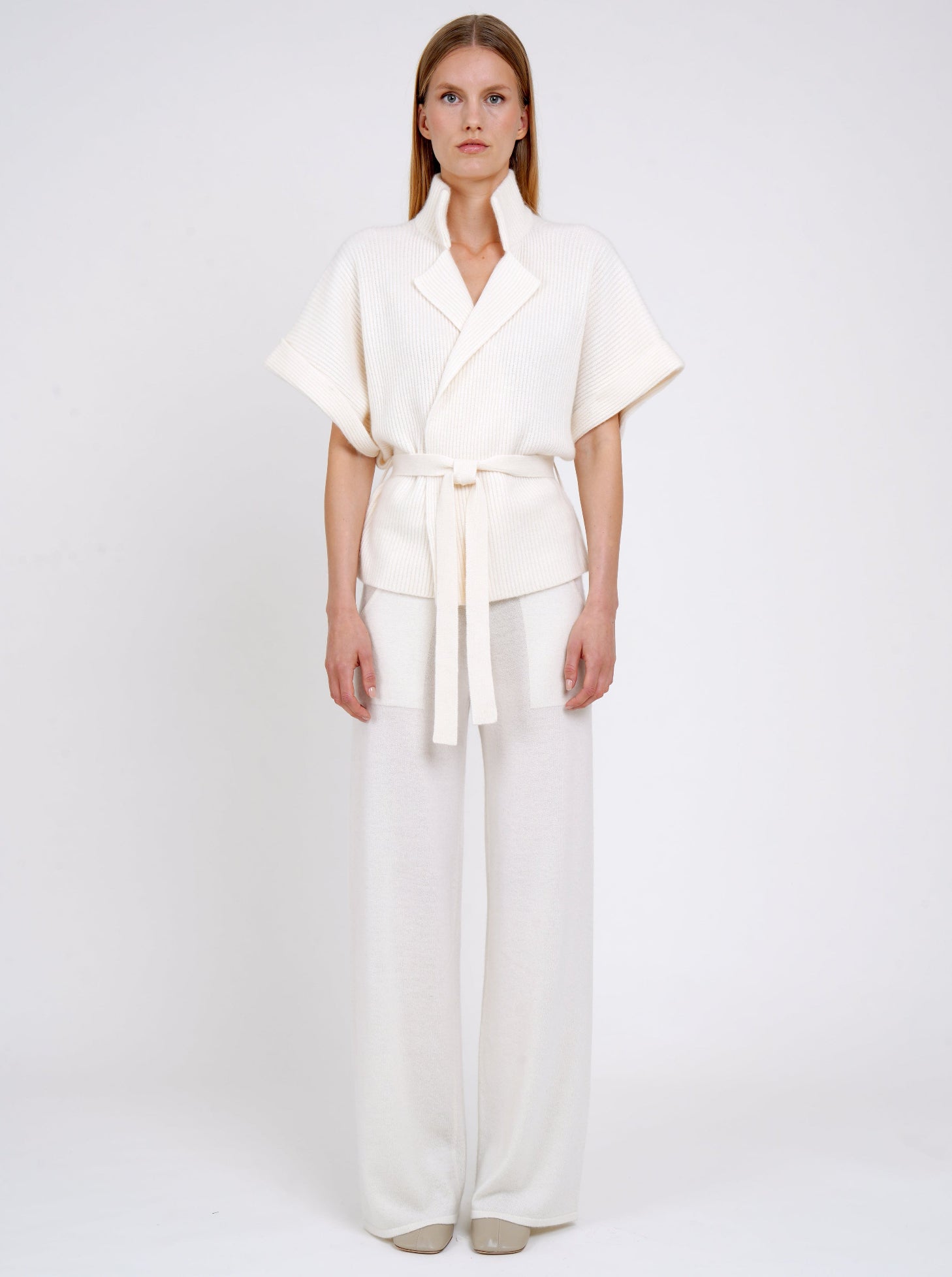 Front view model in white ribbed knit cashmere belted jacket with high collar wide short sleeves and white wide leg knit pants.