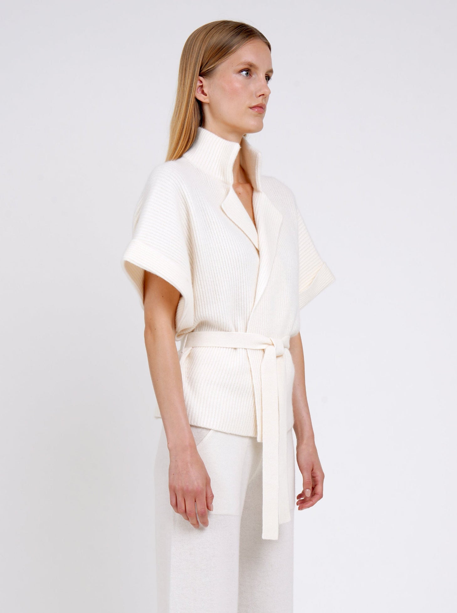 Side view close up model in white ribbed knit cashmere belted jacket with high collar wide short sleeves and white wide leg knit pants.