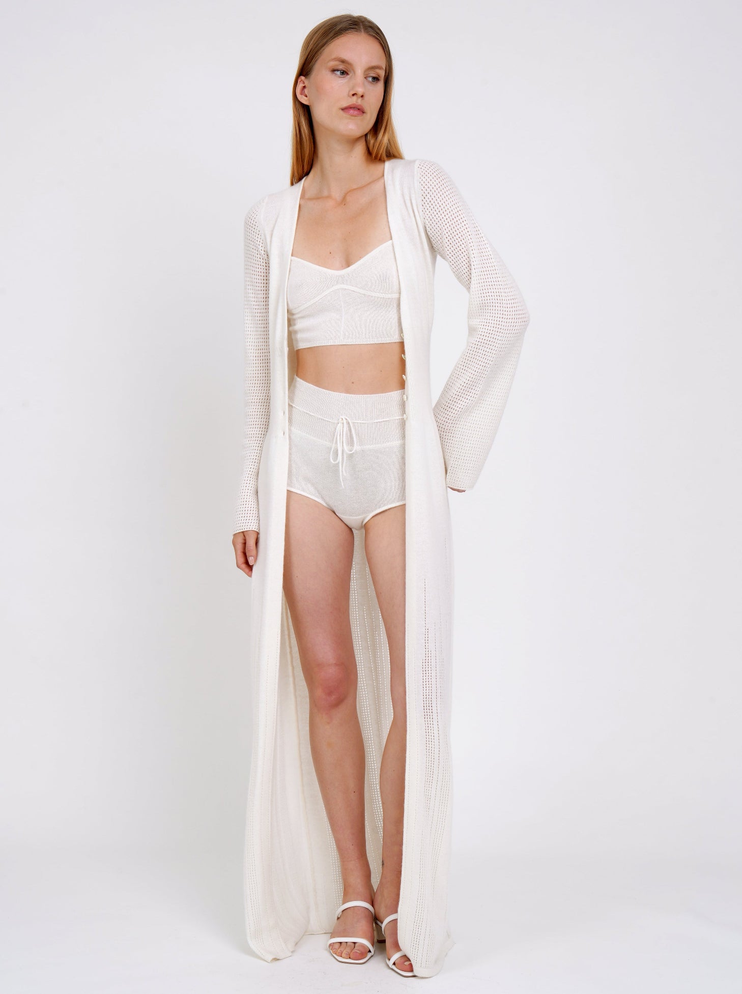 Standing model wears long white knit cardigan over white knit cropped bra top and white cashmere knit matching shorts.