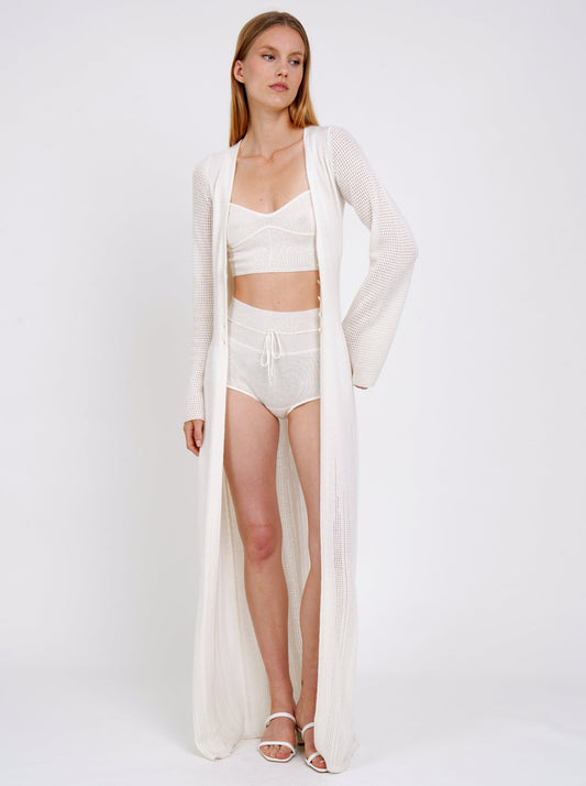 Standing model wears long white knit cardigan over white knit cropped bra top and white cashmere knit matching shorts.