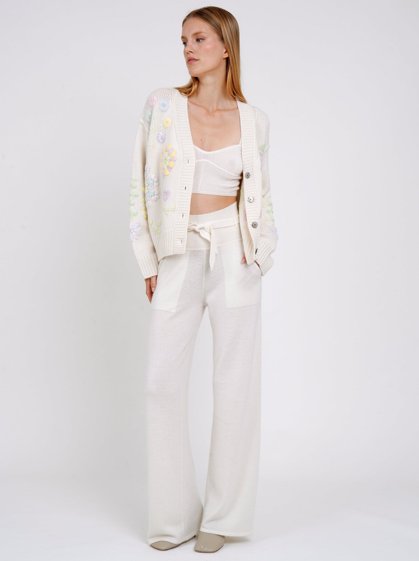 Model wears white embroidered cardigan over white cashmere knit cropped bra top and white cashmere knit high waist trousers.