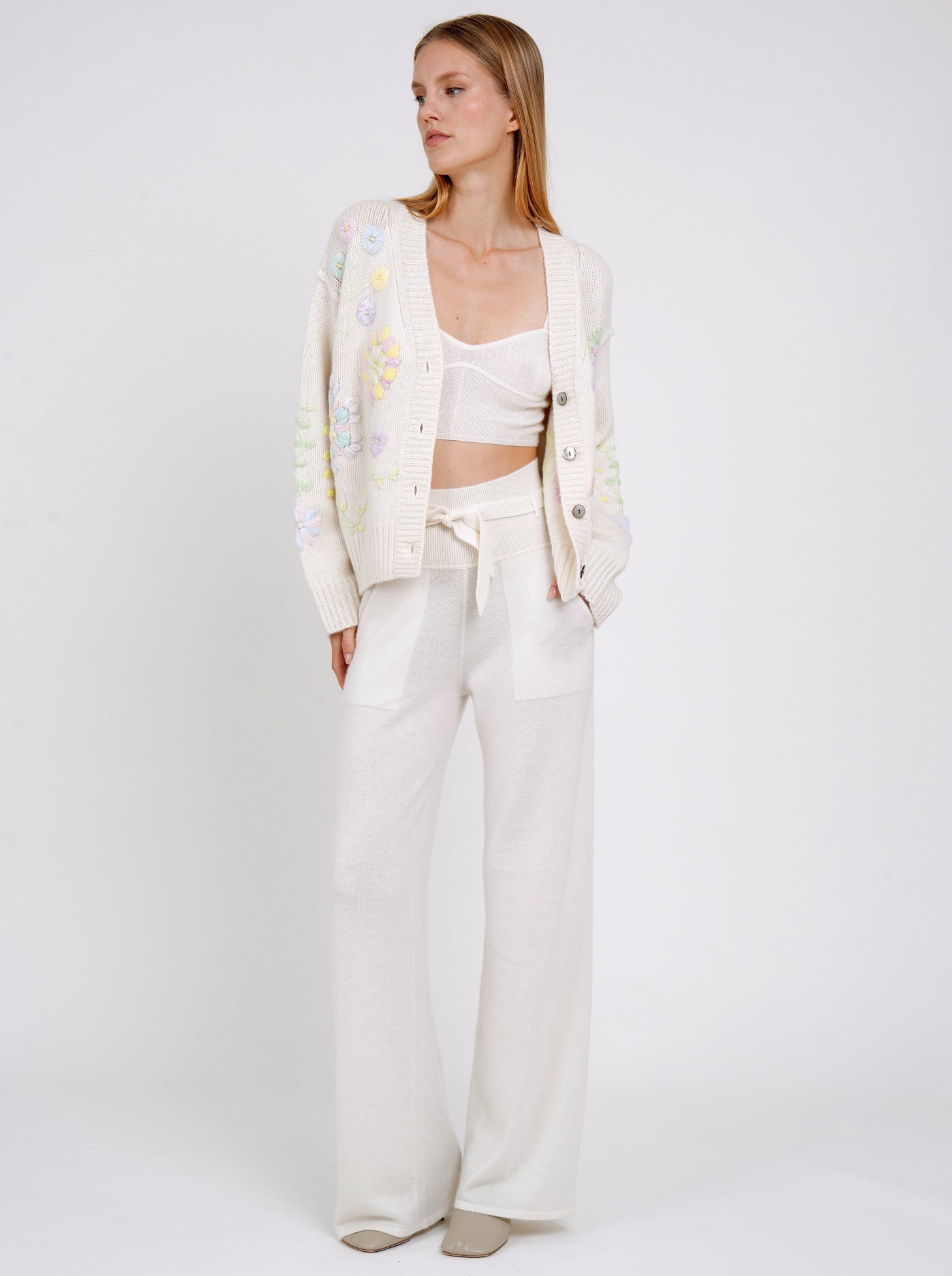 Model wears white embroidered cardigan over white cashmere knit cropped bra top and white cashmere knit high waist trousers.