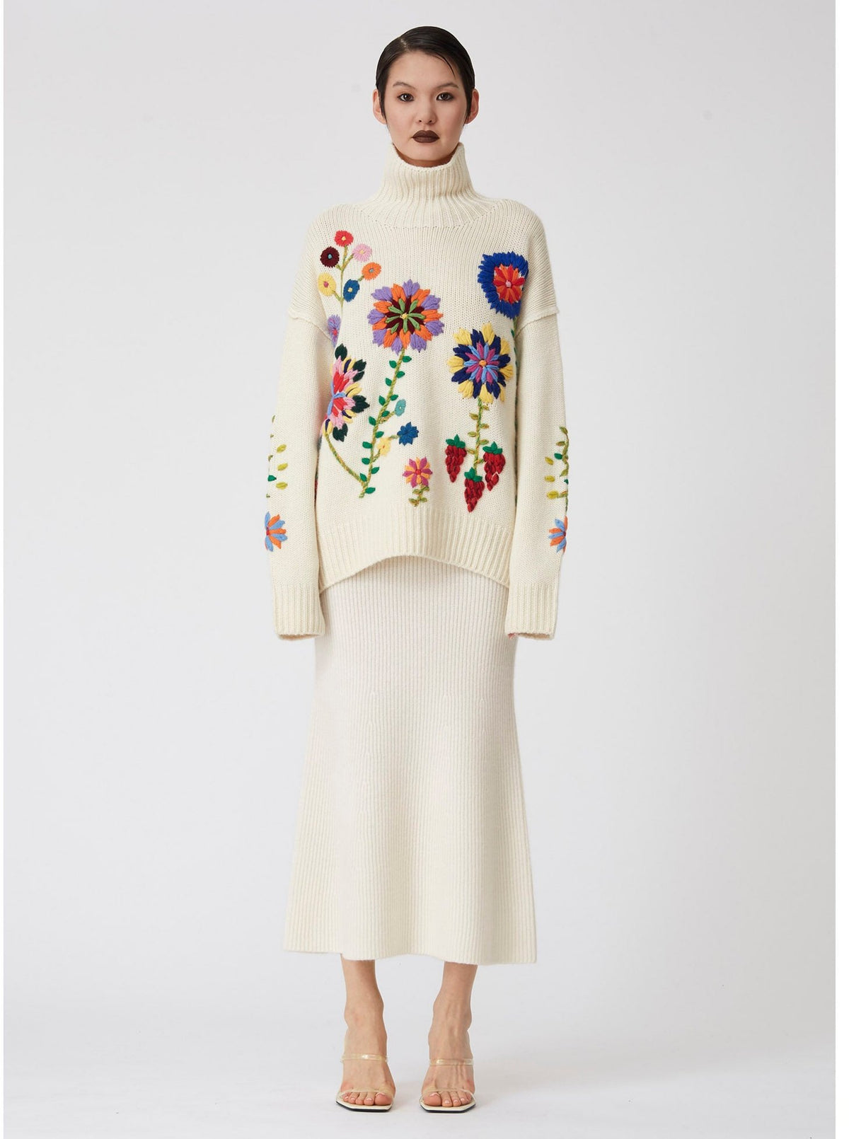 model wear oversized chunky knit white sweater covered in large bright colour hand embroidered flowers; worn with white knit midi skirt and high heels.