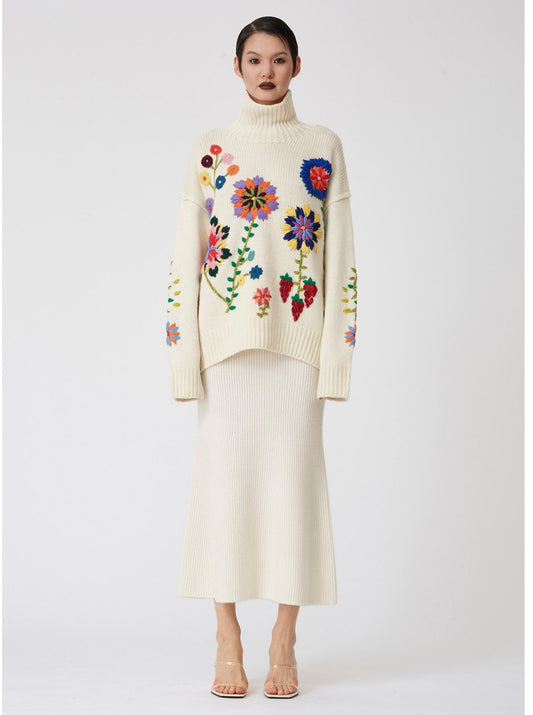 model wear oversized chunky knit white sweater covered in large bright colour hand embroidered flowers; worn with white knit midi skirt and high heels.