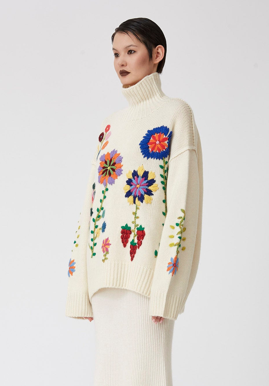 Cropped side view of model wearing oversized chunky knit white sweater covered in large bright colour hand embroidered flowers; worn over white knit midi skirt.