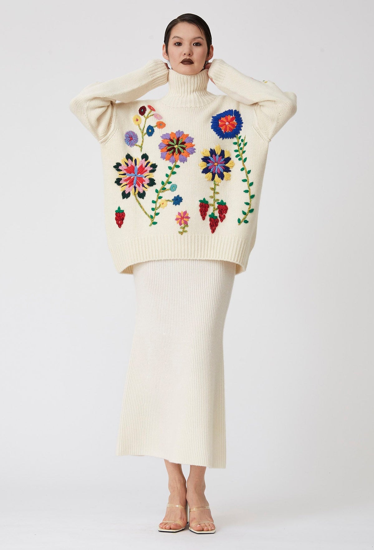 Posed model wearing oversized chunky knit white roll neck sweater covered in large bright colour hand embroidered flowers; worn over white knit midi skirt.